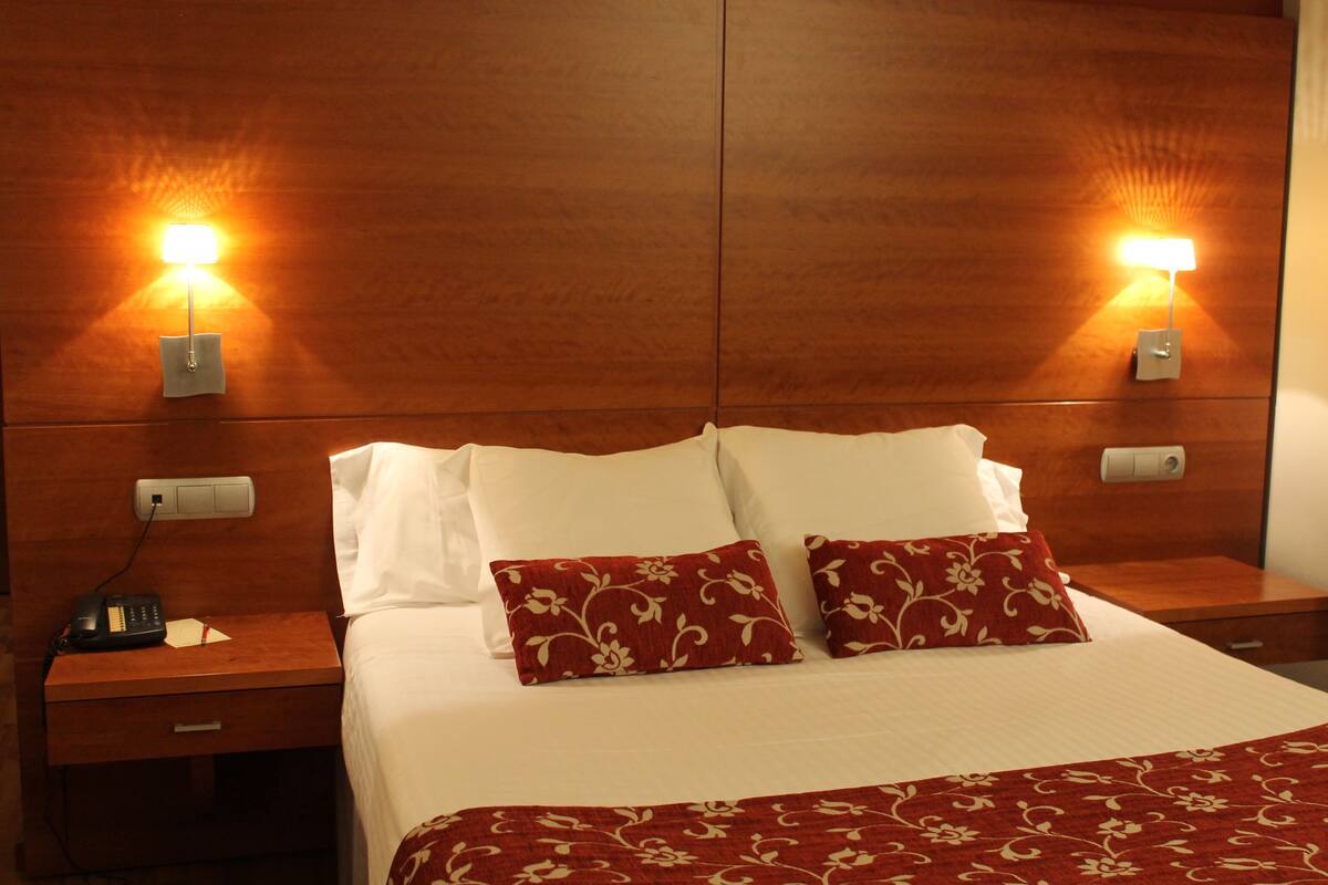 A hotel room with a wooden headboard, two bedside lamps, a telephone, and a bed with white linens and red floral pillows.