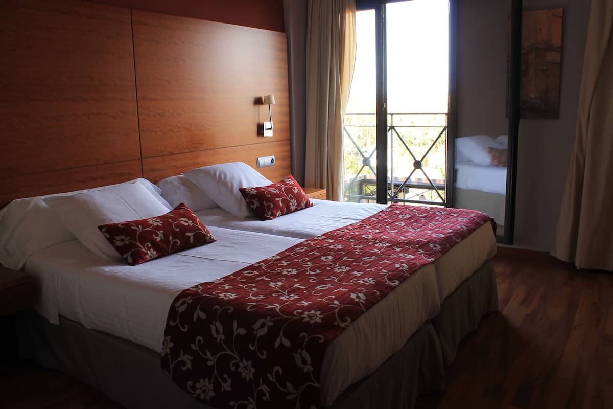 A double bed with a floral bedspread and two pillows, a nightstand, a wall-mounted lamp, and a balcony with a view of a building.