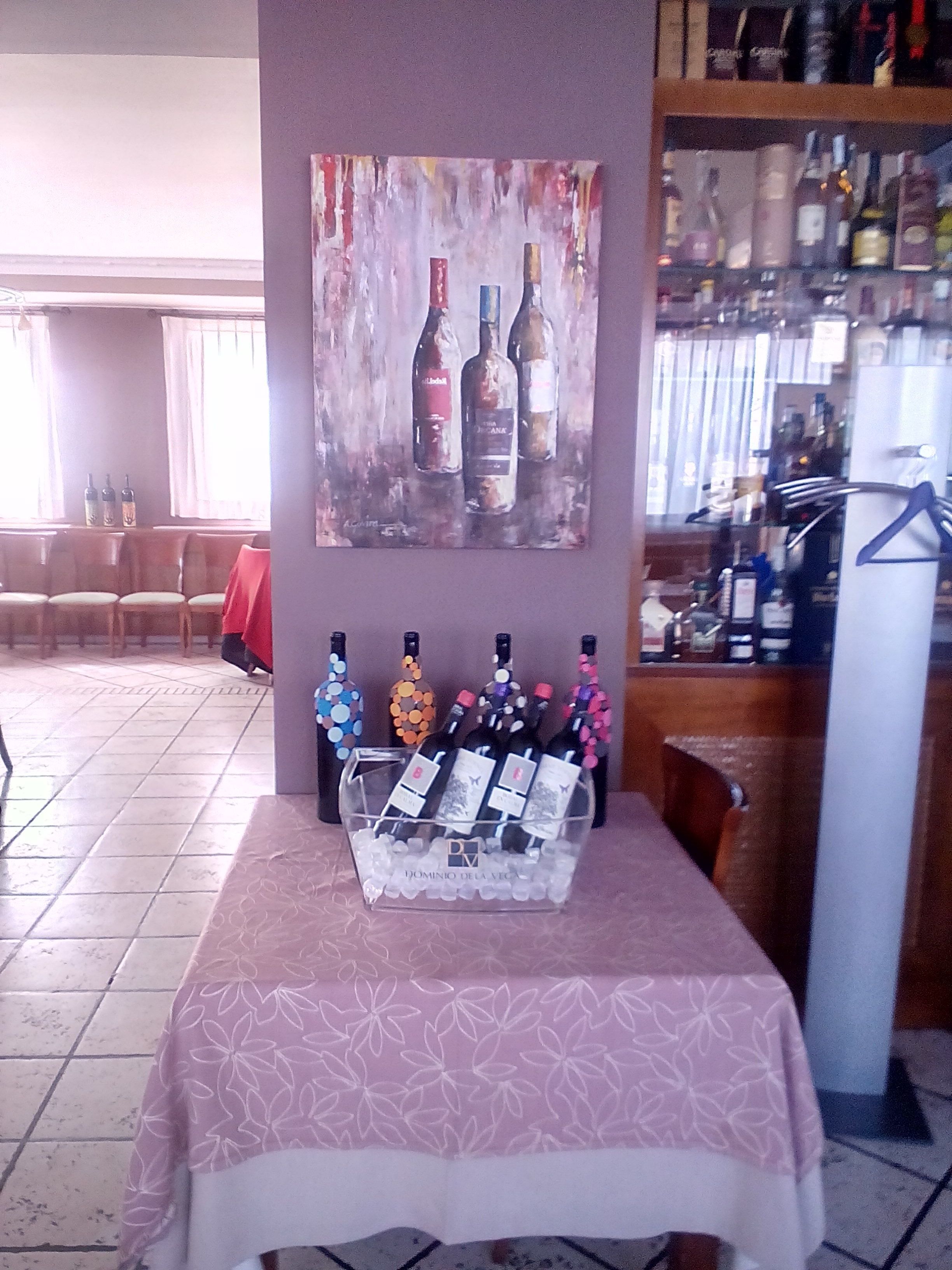 A bar area with a table displaying bottles of wine, a painting of wine bottles on the wall, and a refrigerator.