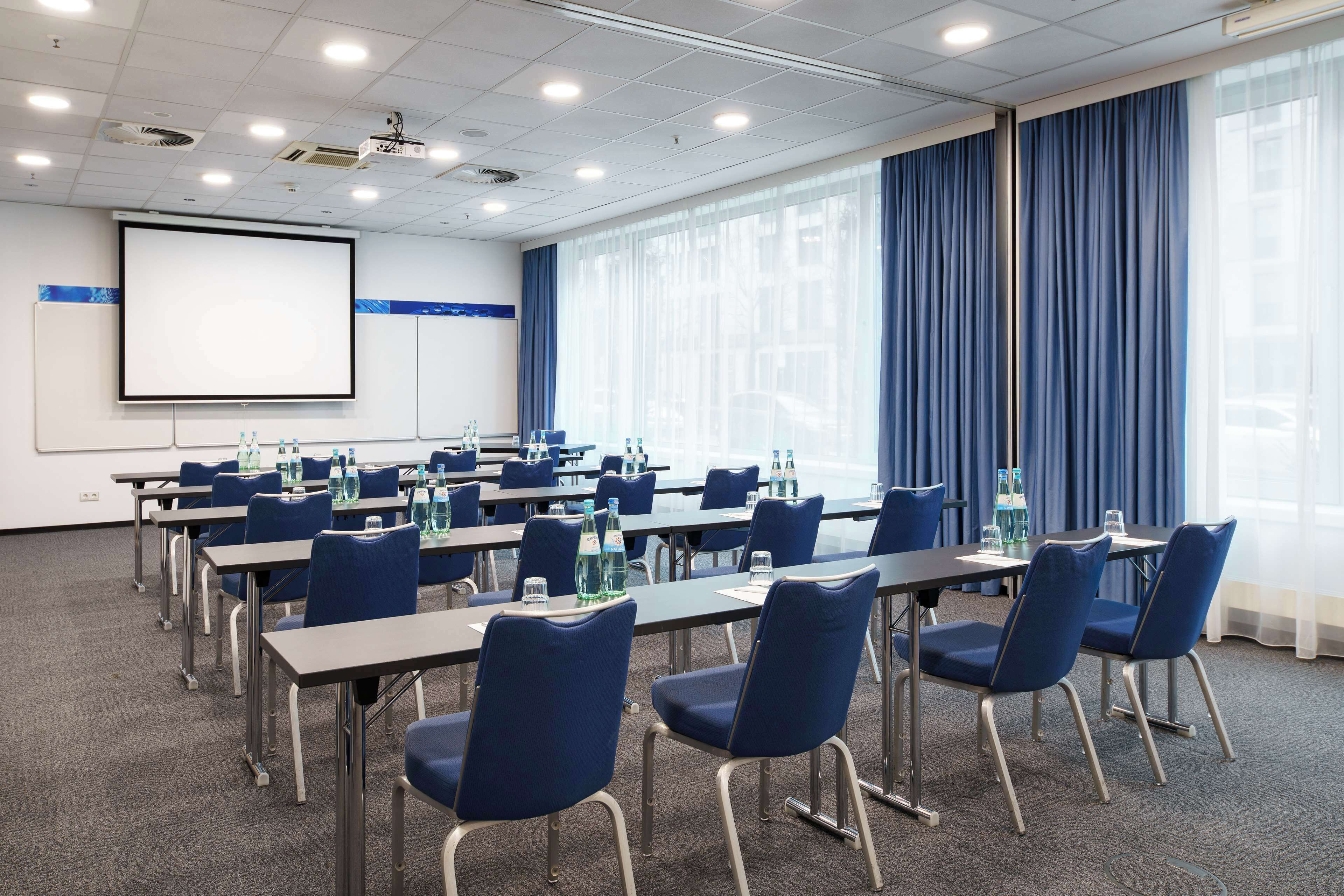 Une salle de conférence équipée de tables, de chaises, d’un écran de projection et d’une grande fenêtre avec des rideaux.