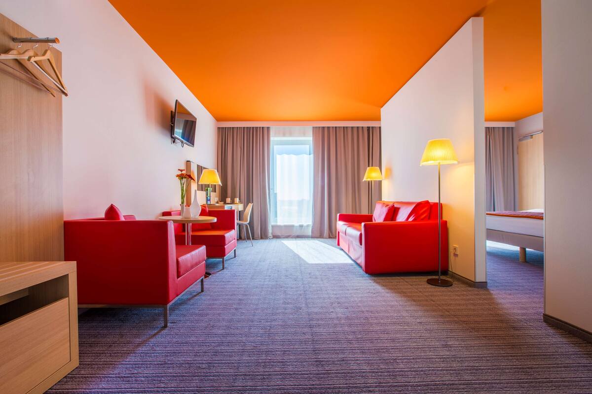 Een hotelkamer met een rode bank, een rode fauteuil, een bureau met een lamp, een aan de muur bevestigde televisie en een groot raam met gordijnen.
