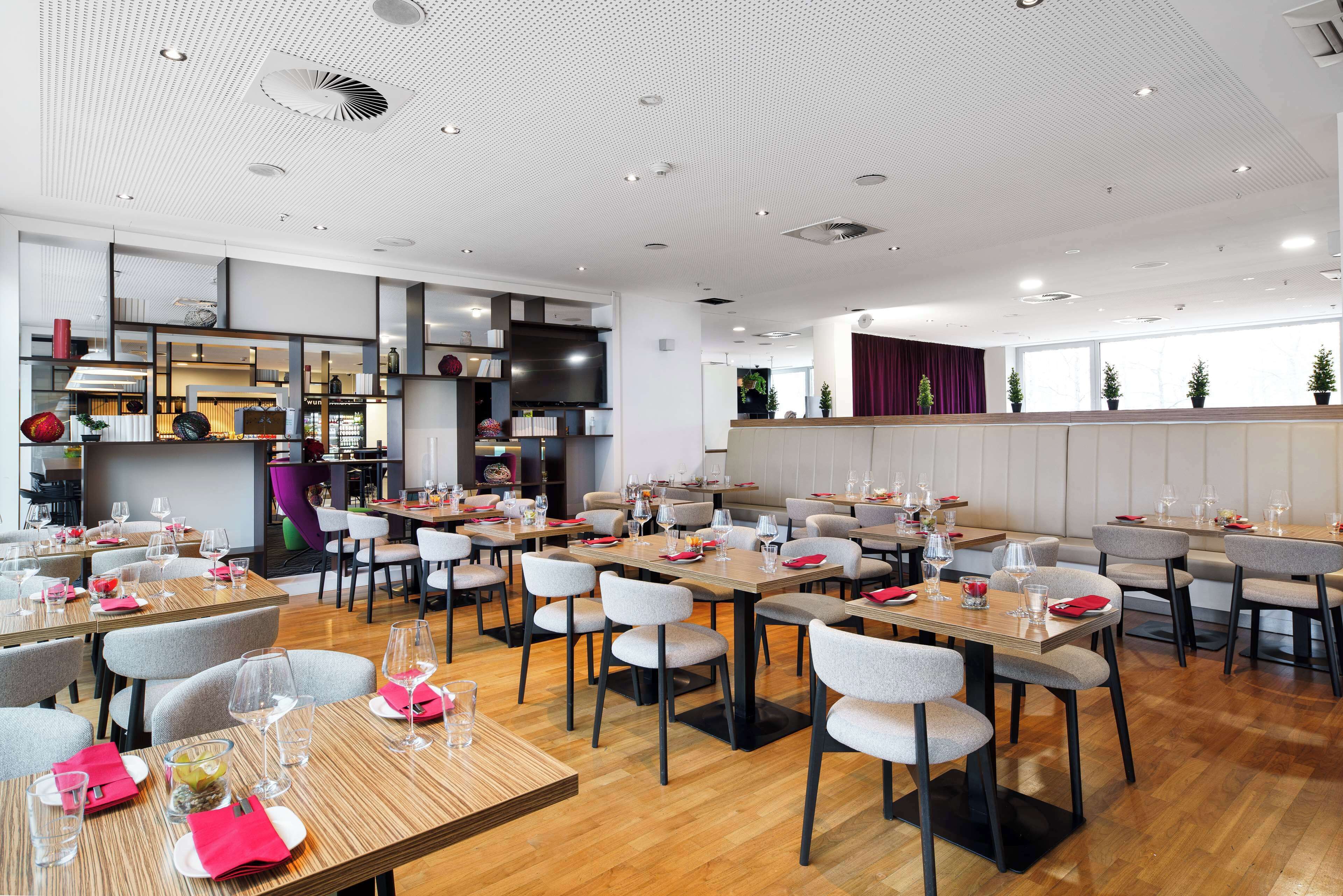 Un restaurante moderno con varias mesas y sillas, una zona de cocina y ventanas amplias.