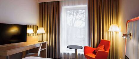 Habitación de hotel con una cama, televisión, escritorio con lámpara, una silla, una mesita y ventana con cortinas.