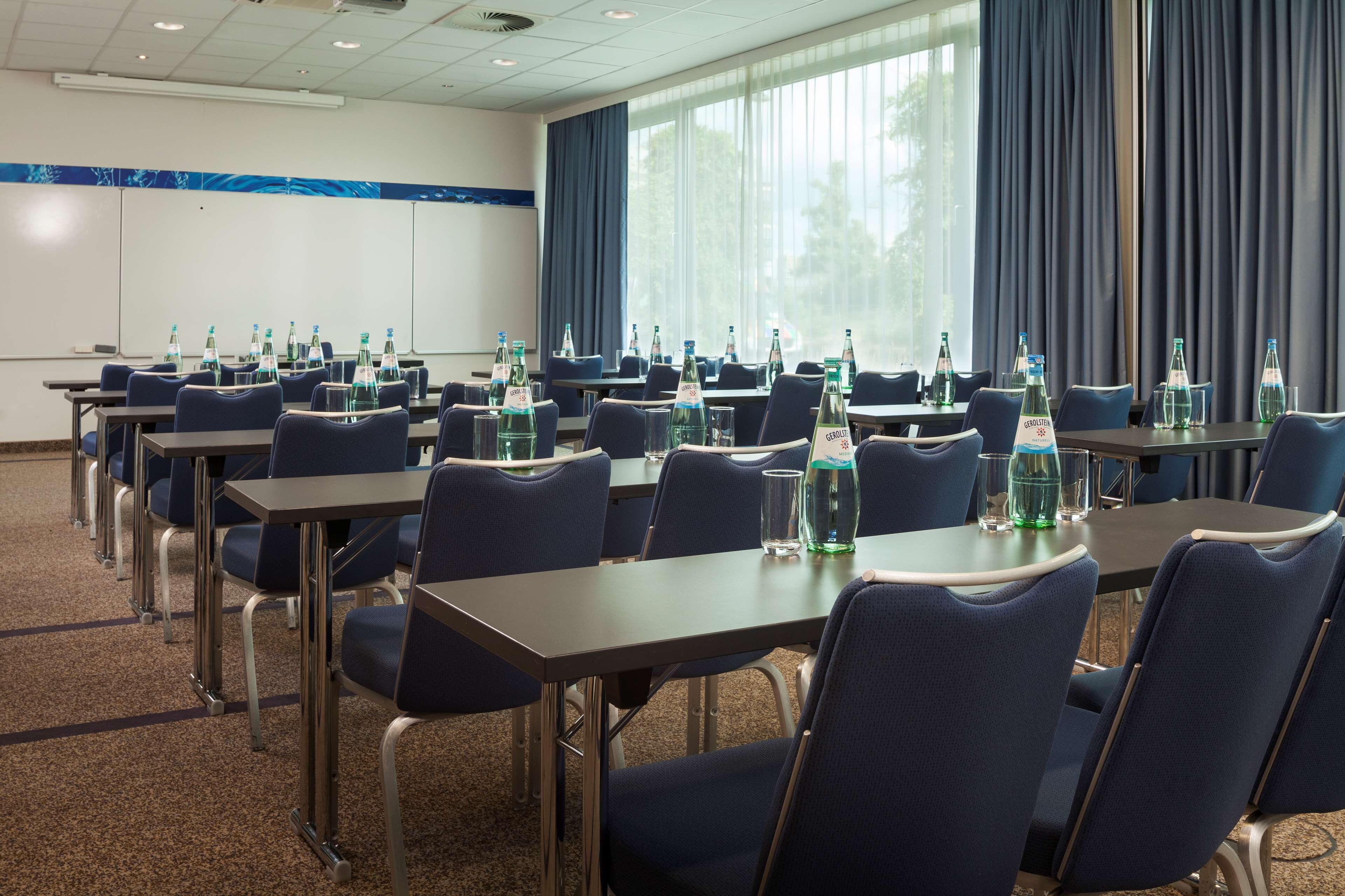 A conference room with multiple tables and chairs, equipped with water bottles and glasses, and large windows with curtains.