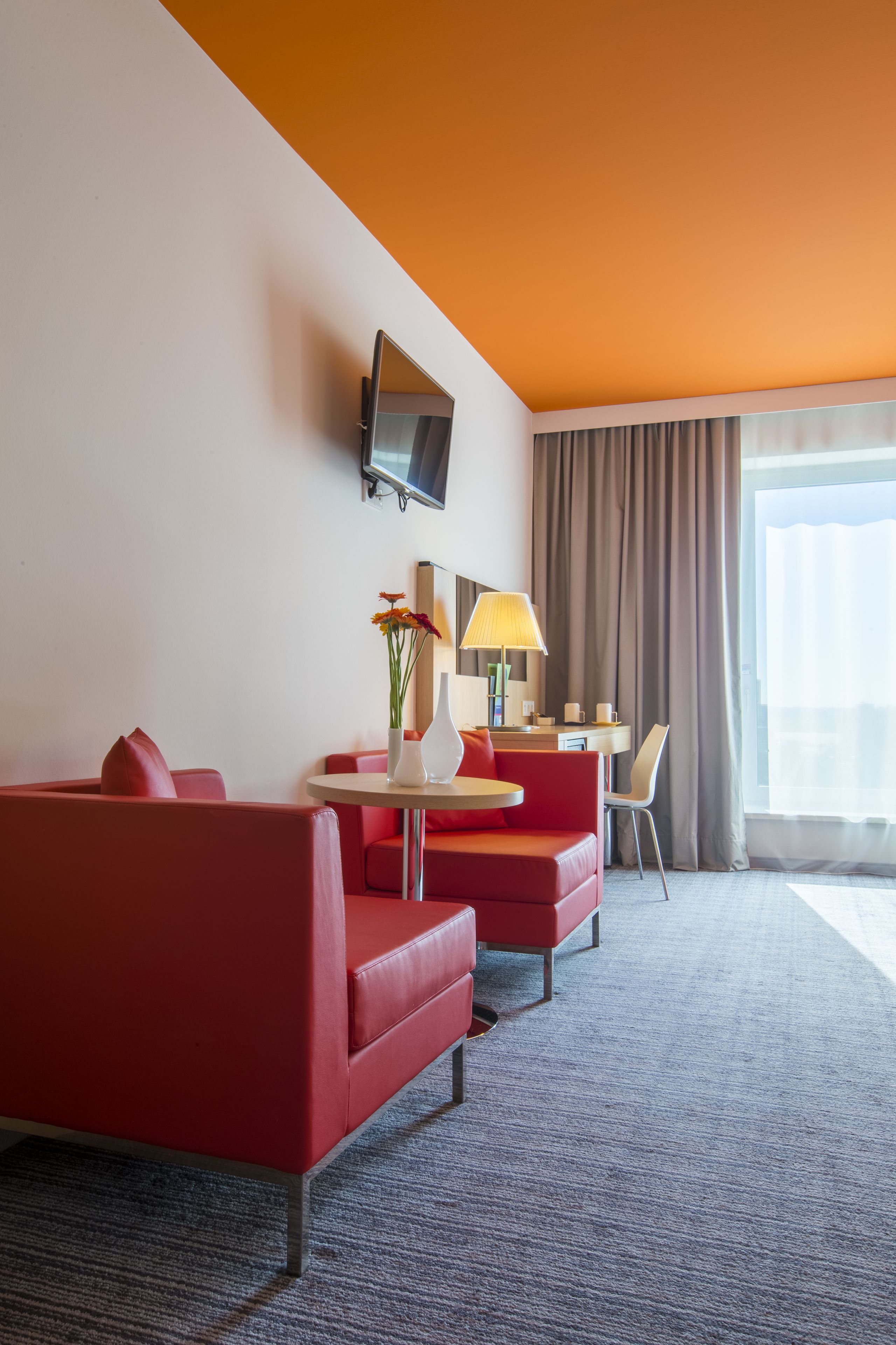 A hotel room with a red sofa, a small table with a vase of flowers, a desk, and a TV mounted on the wall.