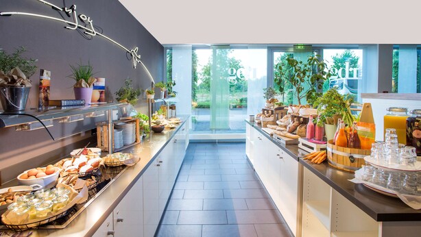 A modern breakfast buffet with a variety of food items and drinks, potted plants, and a large window offering a view of greenery. - Park Inn by Radisson Frankfurt Airport Hotel (Frankfurt)