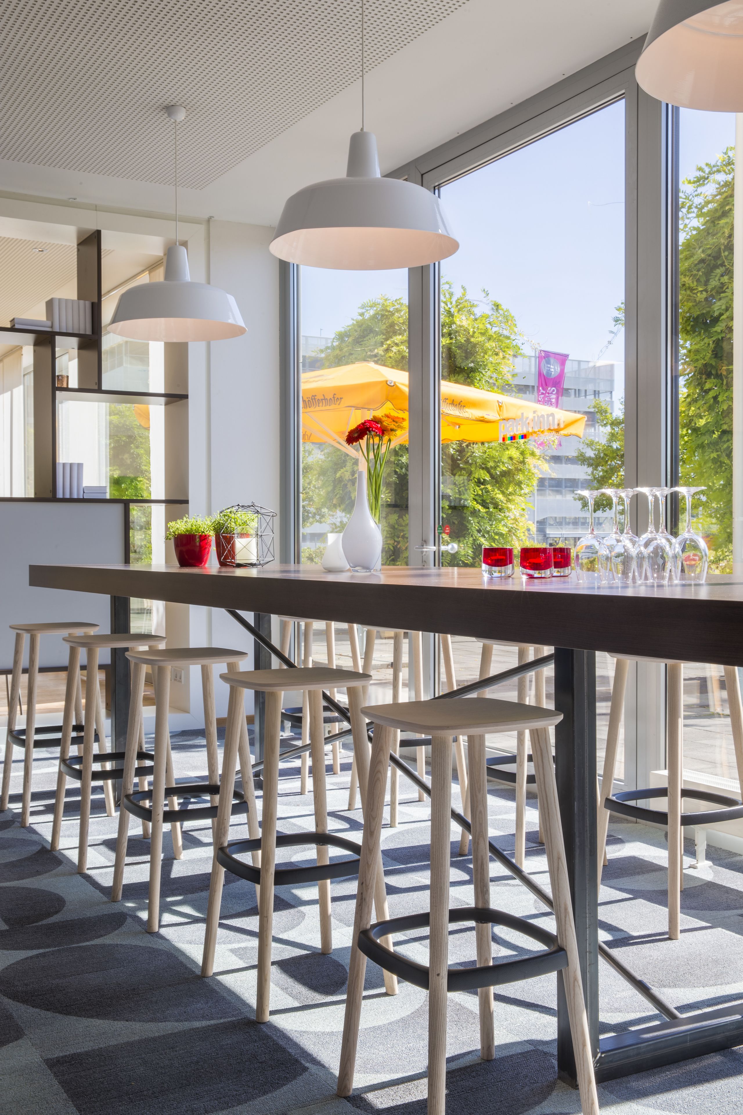 Un área de bar moderna con una barra larga, varios taburetes, lámparas colgantes y amplias ventanas que ofrecen vistas al exterior.