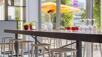 Un área de bar moderna con una barra larga, varios taburetes, lámparas colgantes y amplias ventanas que ofrecen vistas al exterior.