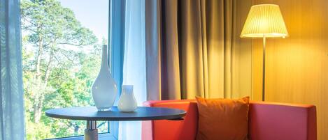 A hotel room with a red sofa, a round table, a lamp, and a view of trees through a window.