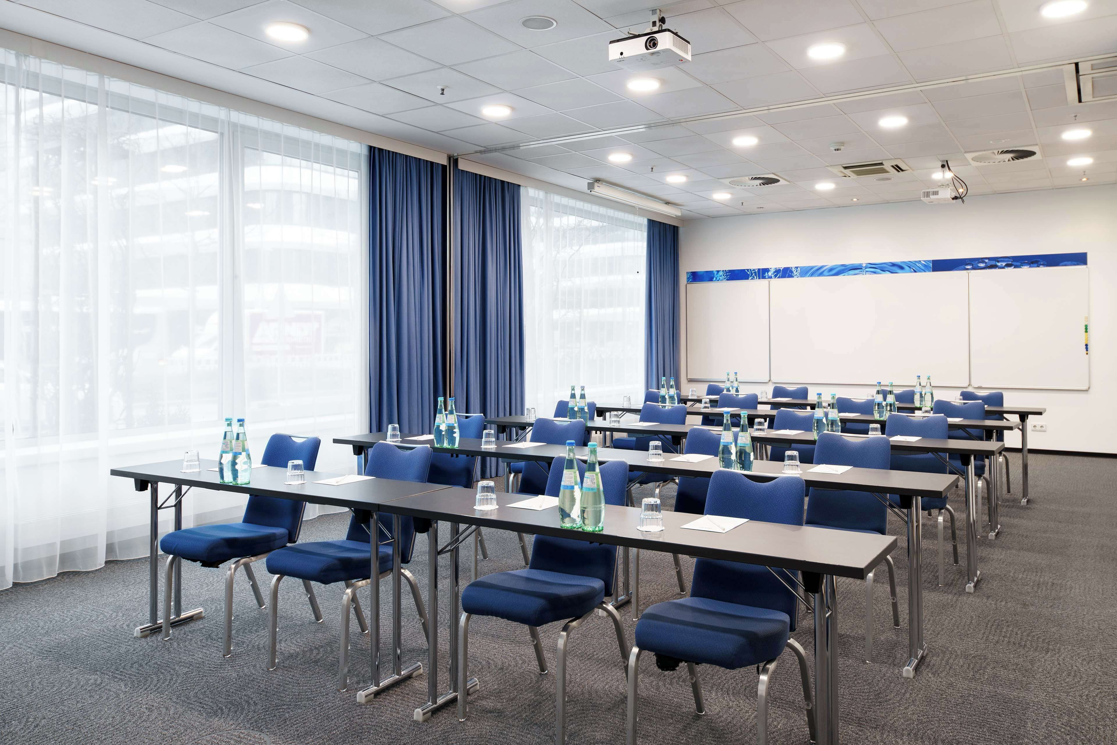 Un salón de conferencias con filas de mesas y sillas, ventanas amplias con cortinas azules y una pizarra en la pared.