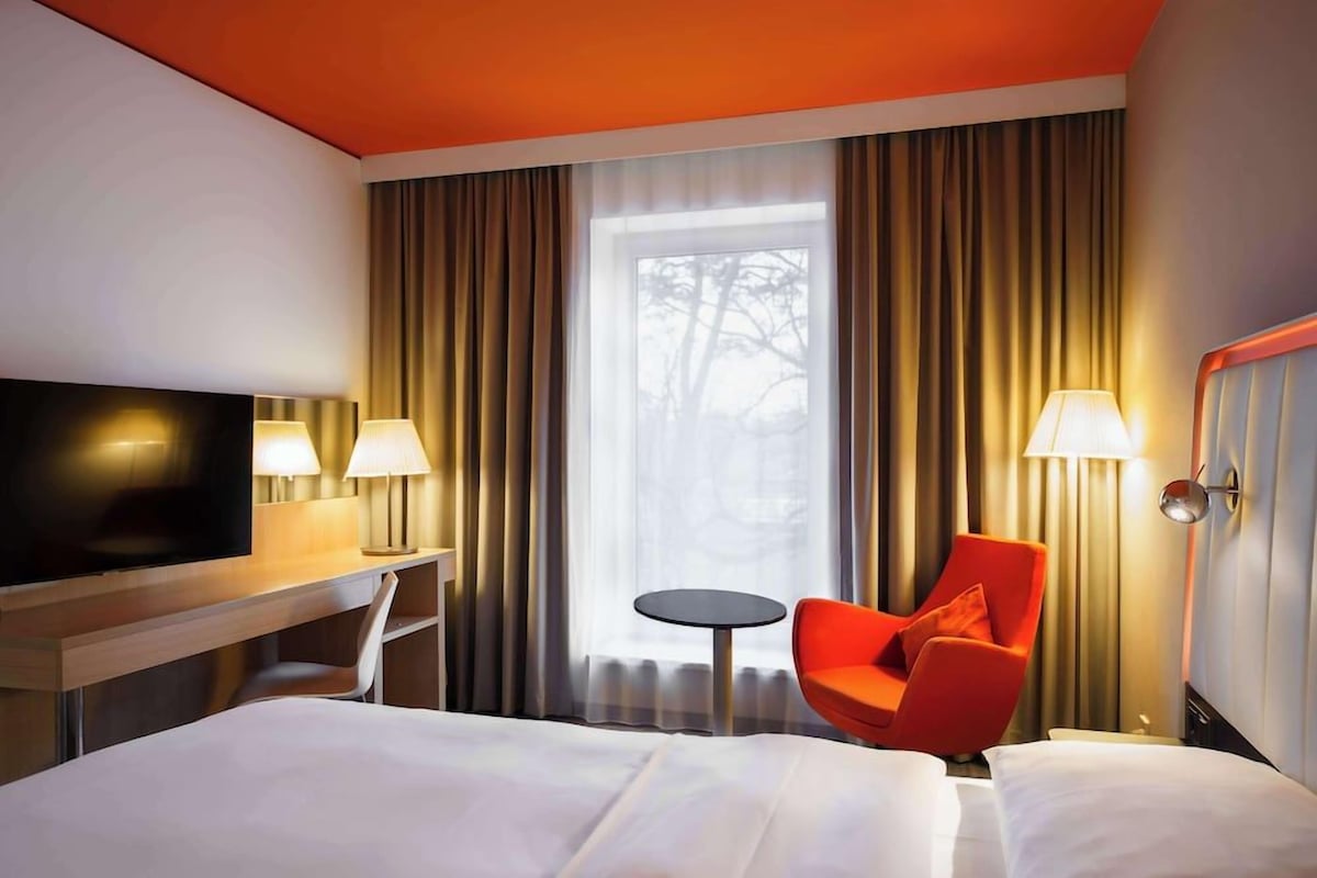 A hotel room with a bed, a TV, a desk with a lamp, a chair, a small table, and a window with curtains.