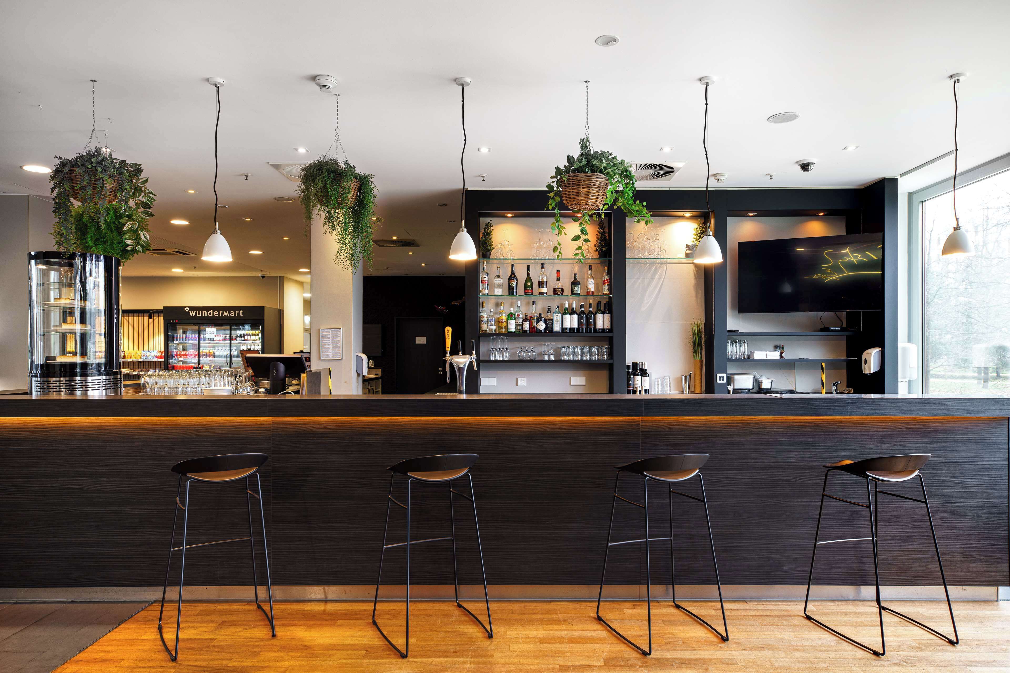 A modern bar area with a wooden counter, three bar stools, hanging plants, a television, and a shelf with bottles.