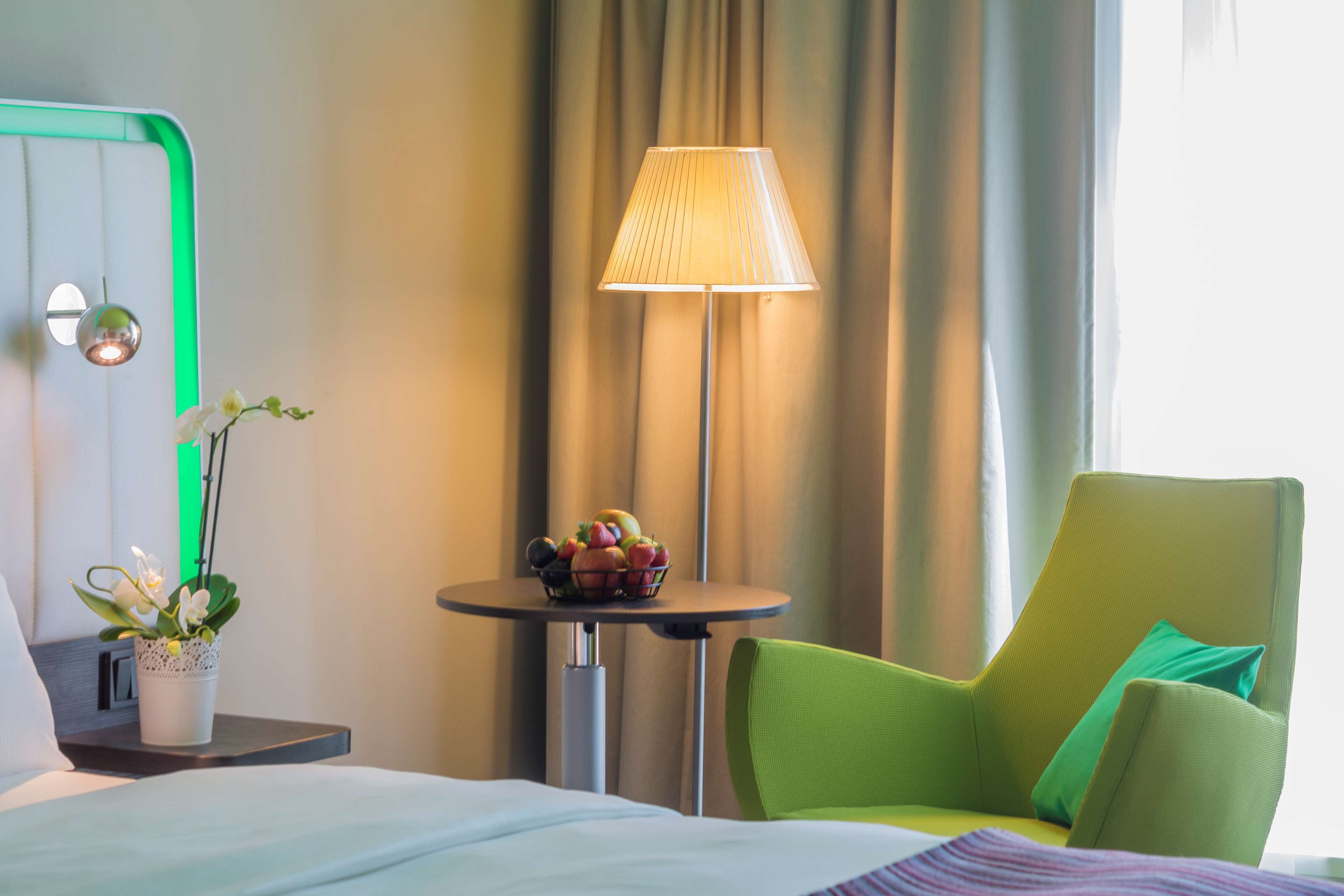 Habitación de hotel con cama, un sillón verde, una mesita con fruta, una lámpara y una planta en maceta.