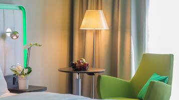 A hotel room with a bed, a green armchair, a small table with fruit, a lamp, and a potted plant.