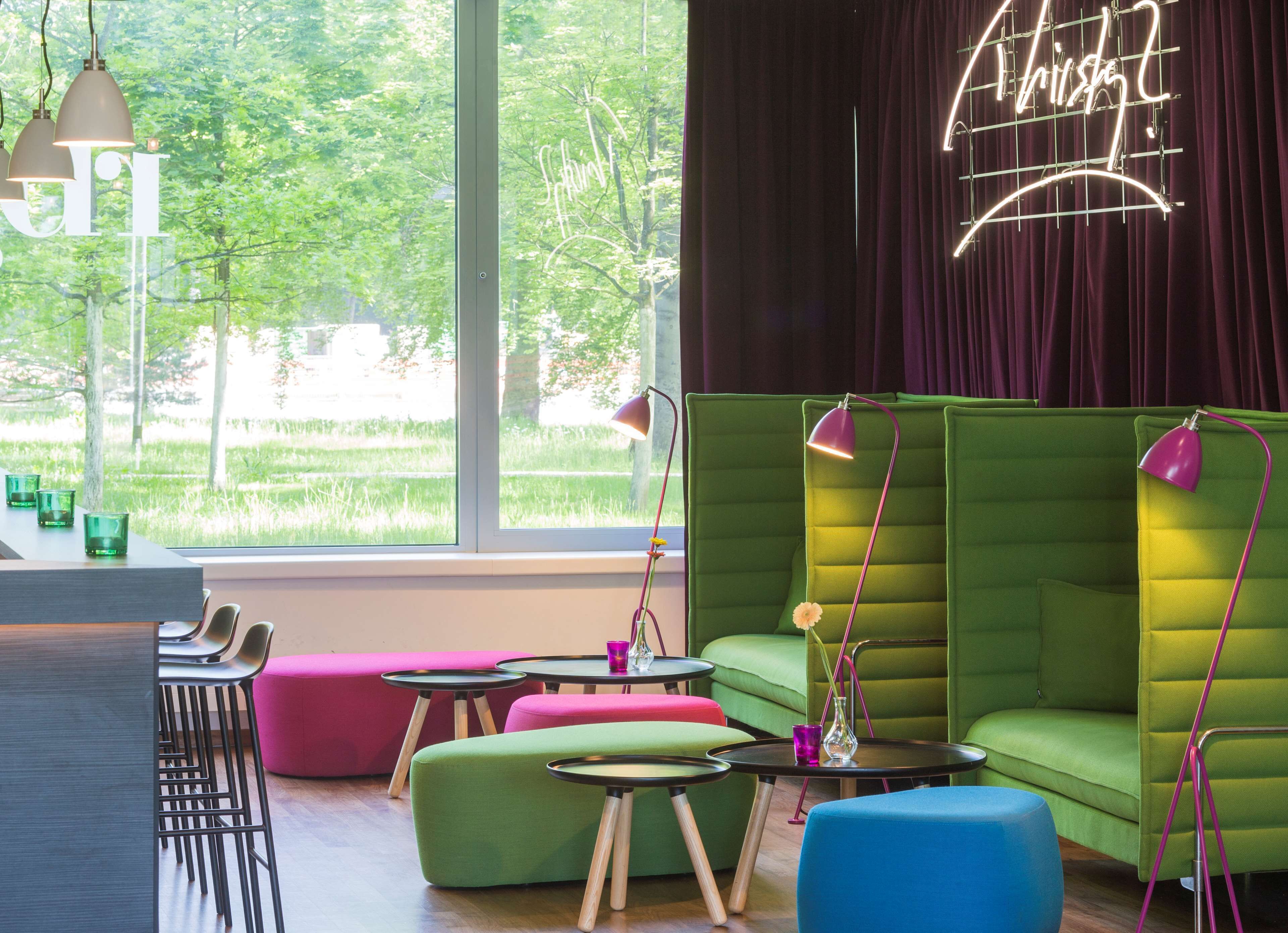 Eine moderne Lounge mit grünen Sofas, einem großen Fenster mit Blick ins Grüne und einem Neonzeichen an der Wand.