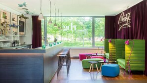A modern bar area with a counter, stools, and a bar stocked with bottles. There are colorful seating areas with green and pink couches and a large window offering a view of greenery. - Park Inn by Radisson Frankfurt Airport Hotel (Frankfurt)