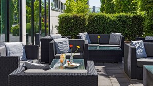A patio area with wicker furniture, including sofas and chairs, arranged around a glass-top table with a plate of food and drinks. - Park Inn by Radisson Frankfurt Airport Hotel (Frankfurt)
