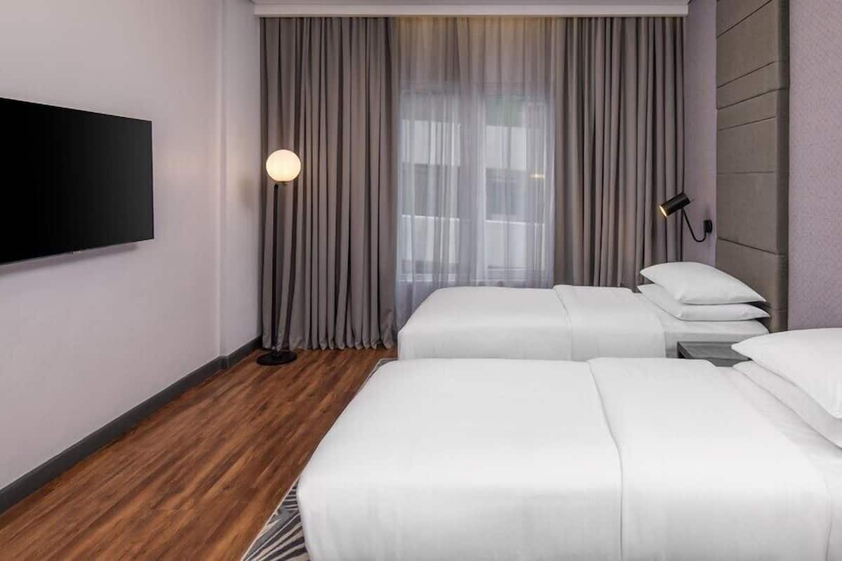 A hotel room with two beds, a flat-screen TV mounted on the wall, a floor lamp, and a window with sheer curtains.