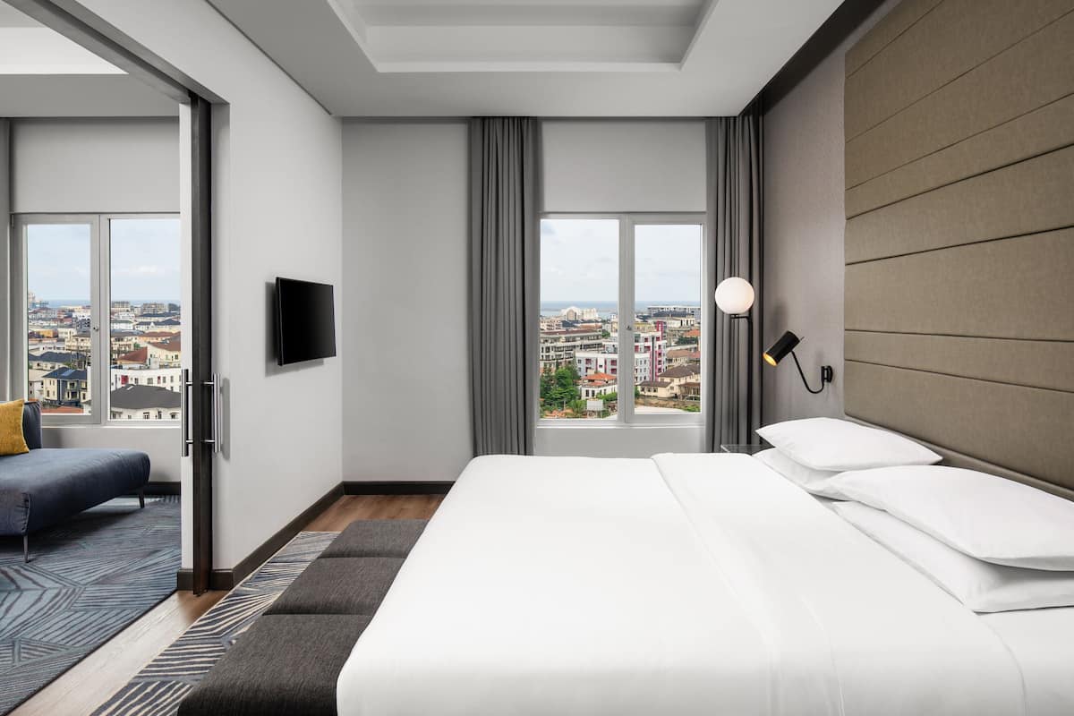 A modern hotel room with a large bed, a TV, and a city view through the windows.