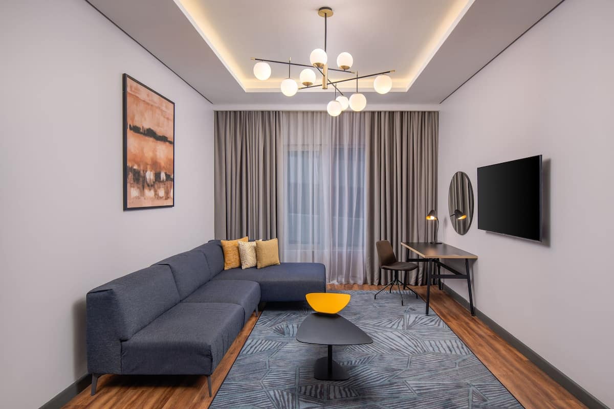 A modern living room with a sectional sofa, a small table with a yellow bowl, a TV mounted on the wall, and a dining area with a table and chairs.