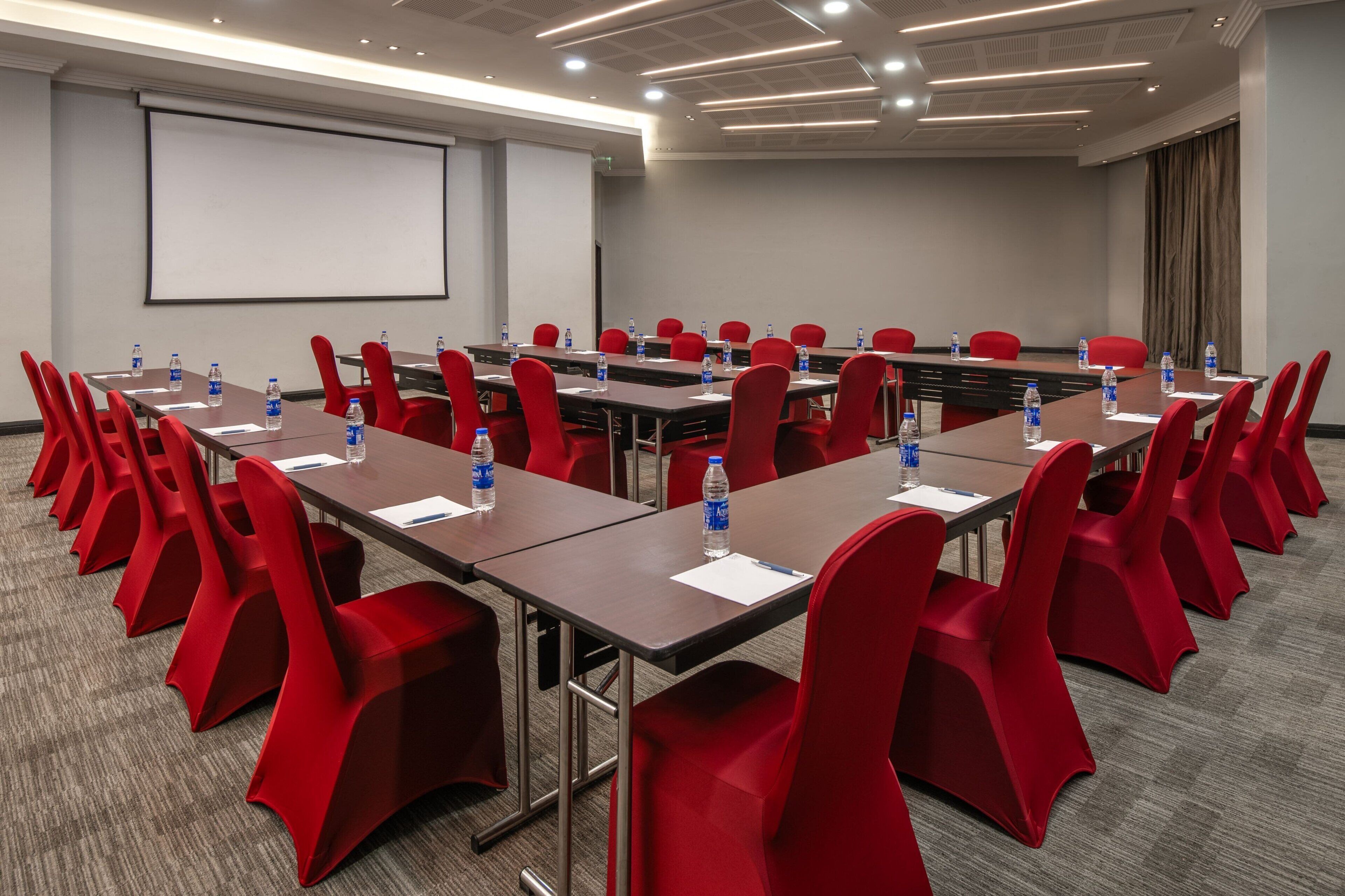 A conference room with rows of red chairs, tables with water bottles, a projector screen, and a carpeted floor.