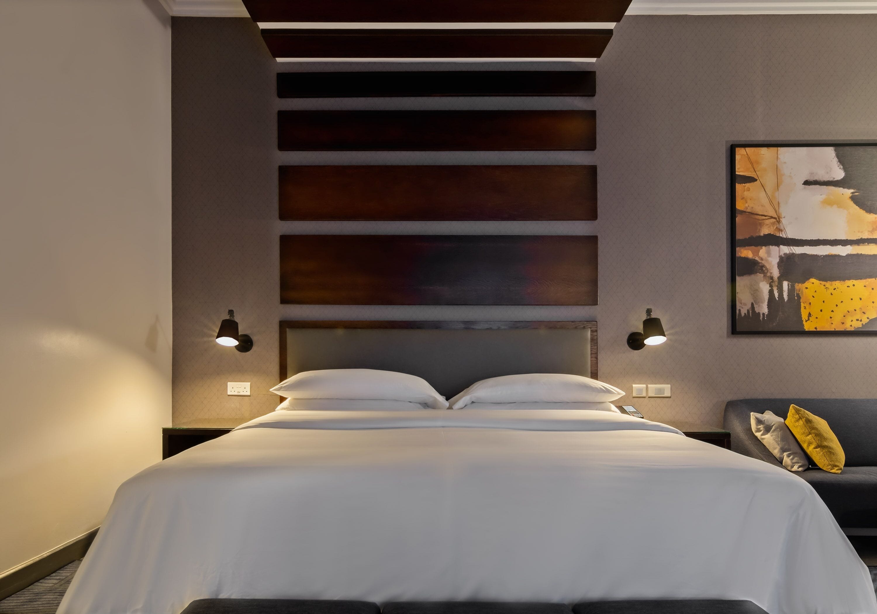 Ett modernt hotellrum med en stor säng, en sänggavel med horisontella paneler, två väggmonterade lampor och en tavla på väggen.