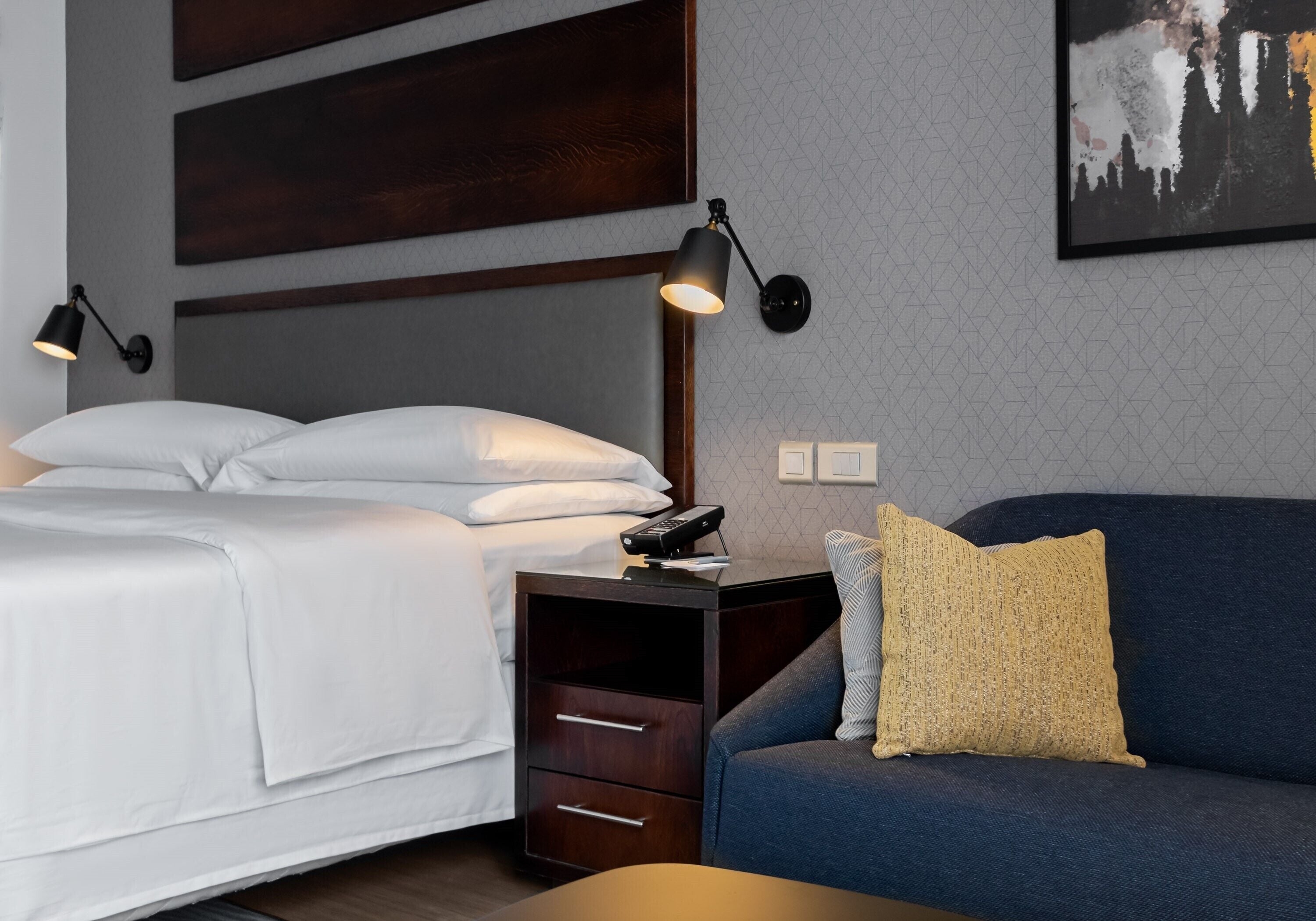 Une chambre d’hôtel avec un lit, une table de chevet, une lampe et un canapé avec un coussin.