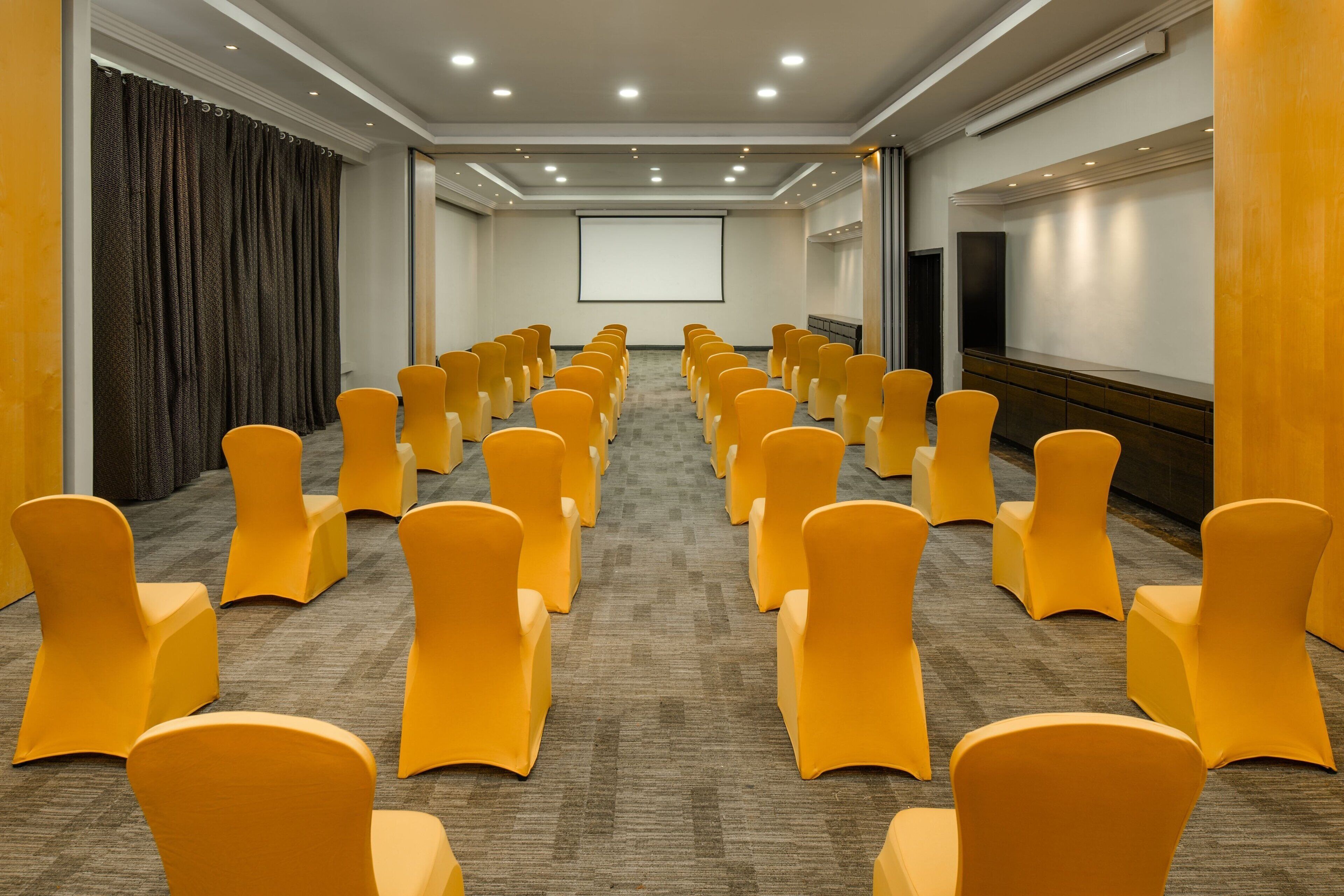 Une salle de conférence avec des rangées de chaises jaunes, un grand écran et un sol recouvert de moquette.