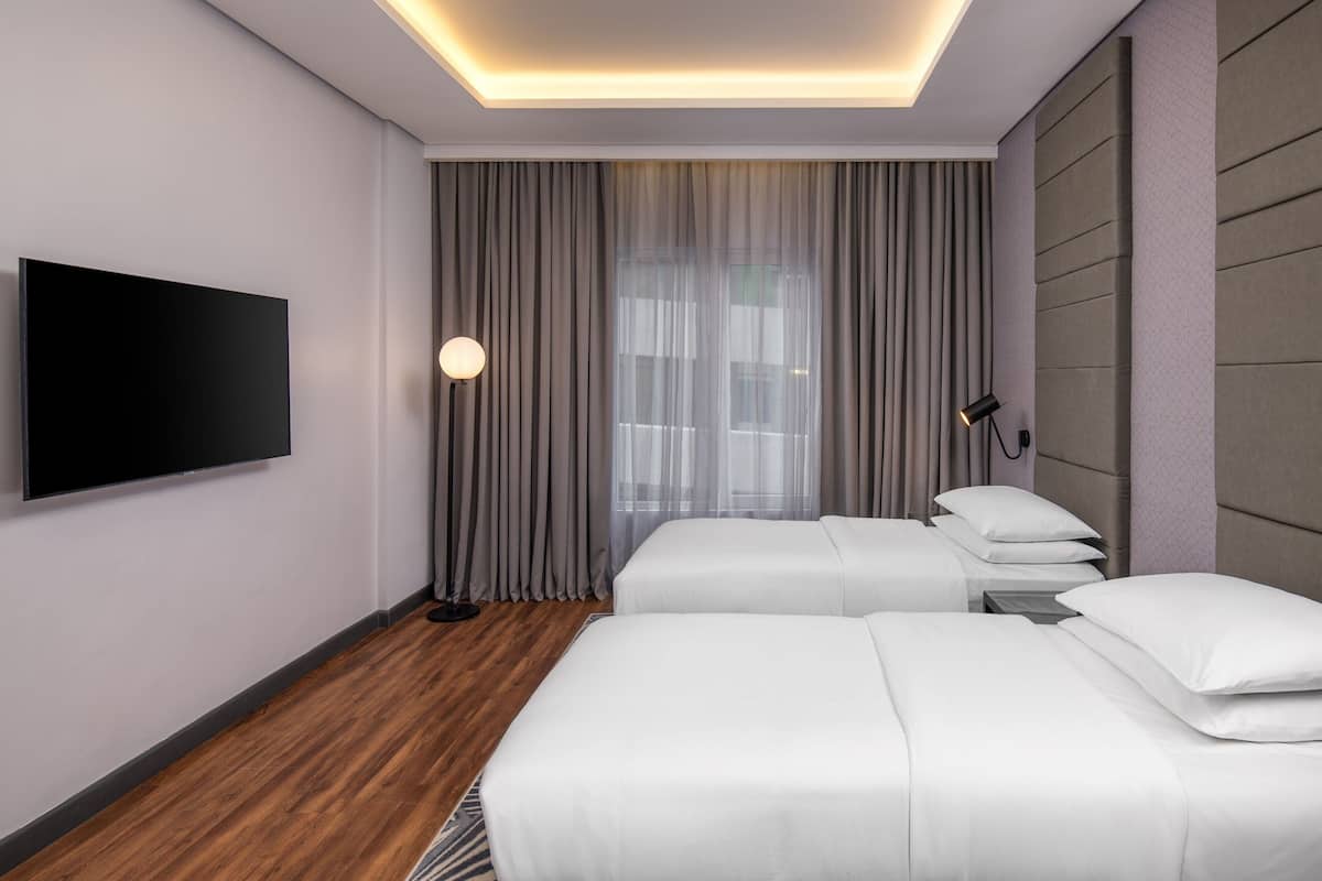 A hotel room with two beds, a flat-screen TV, a bedside lamp, and a large window with curtains.