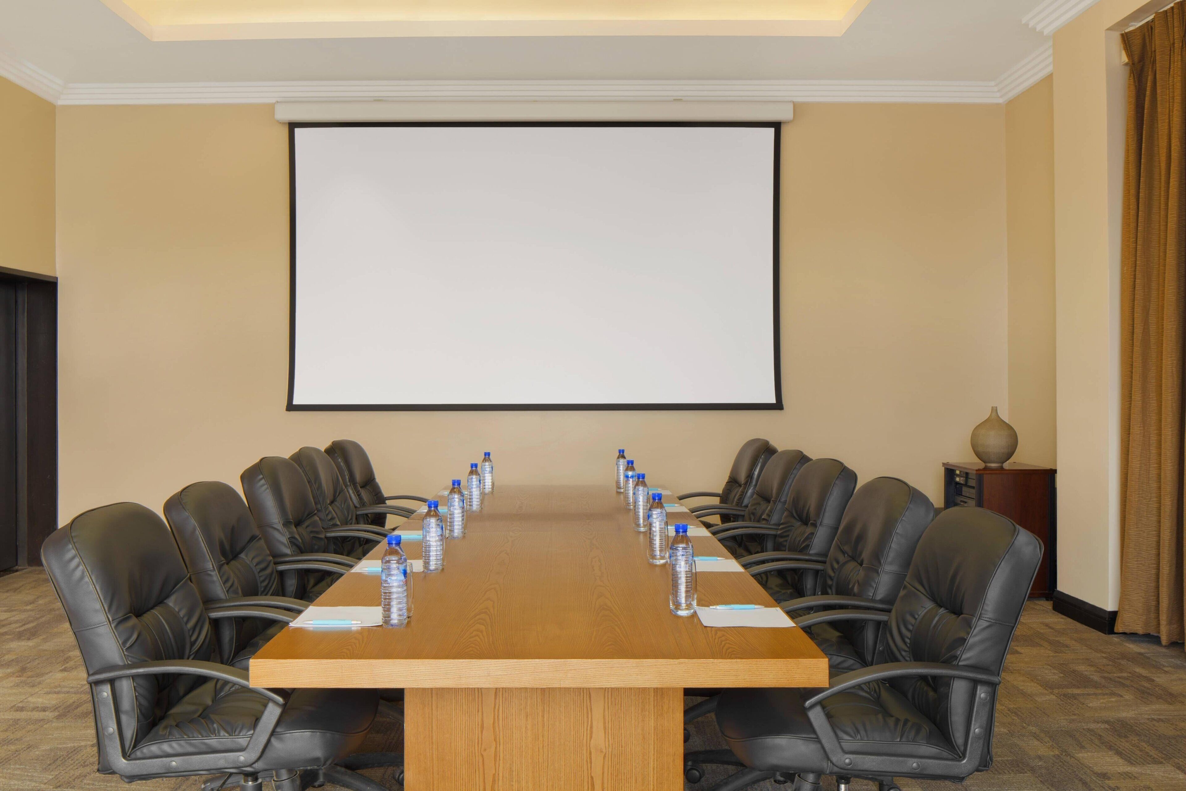 Un salón de conferencias con una larga mesa, sillas de cuero negro, botellas de agua y una pantalla de proyector.