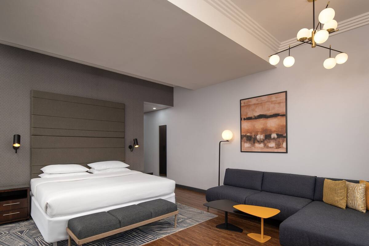 A modern hotel room with a large bed, a sofa, a small table, and a painting on the wall.
