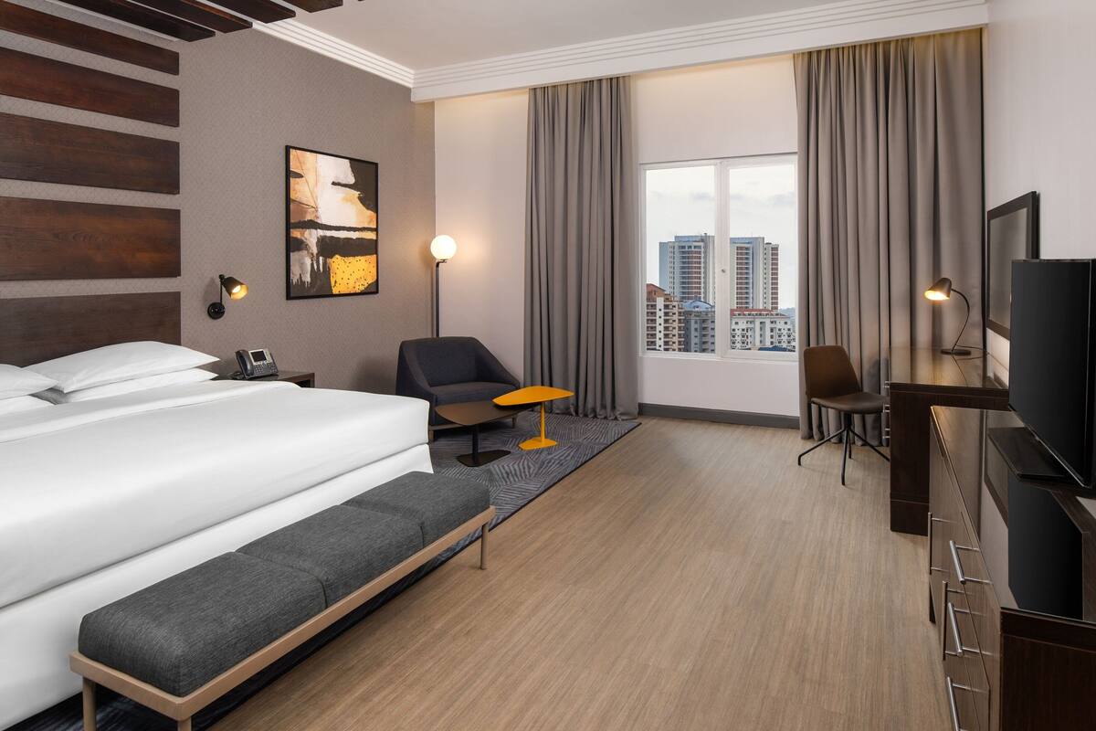 A hotel room with a large bed, a desk with a TV, a chair, and a view of the city.