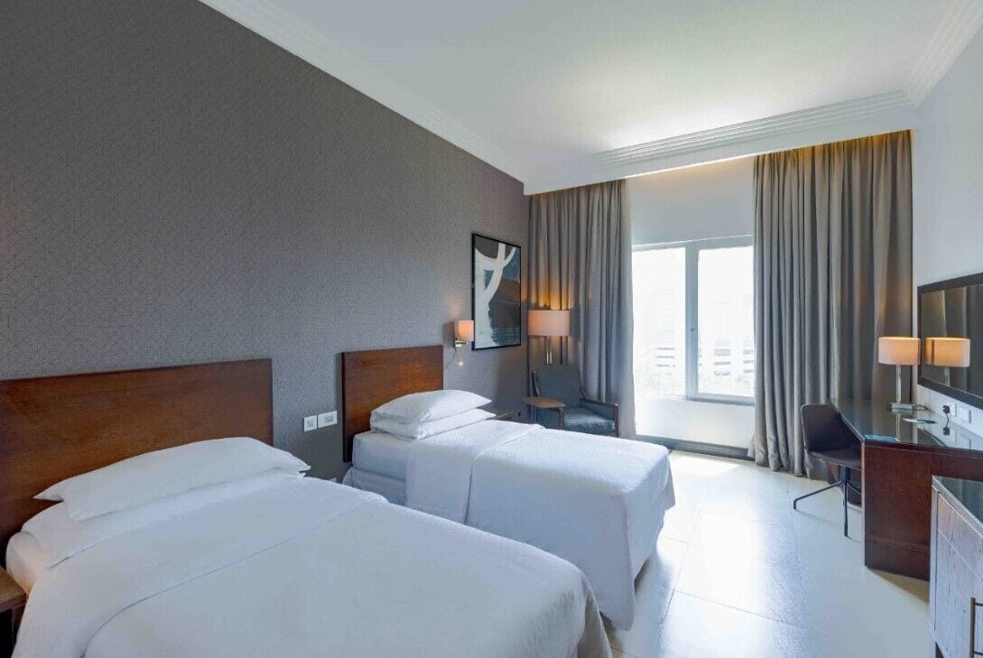 A hotel room with two beds, a desk, a chair, a lamp, and a large window with curtains.