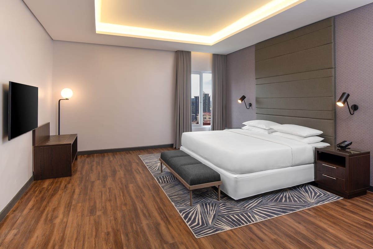 A modern hotel room with a large bed, a flat-screen TV, a bedside table, a nightstand, and a view of the city.