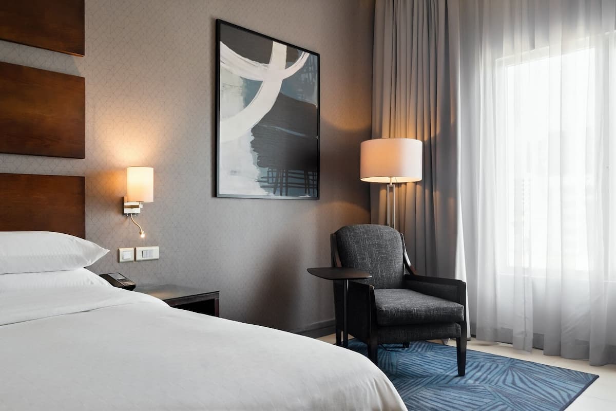 A hotel room with a large bed, a chair, a desk, a lamp, and a painting on the wall.