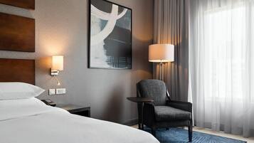 Ett hotellrum med en stor säng, en stol, ett skrivbord, en lampa och en tavla på väggen.