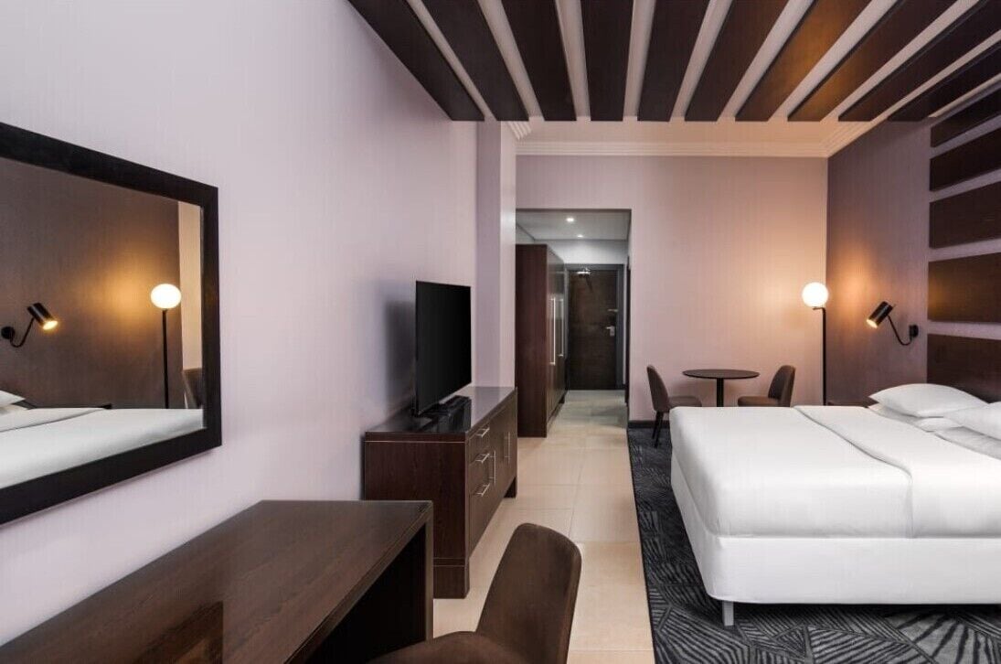 A hotel room with a bed, a desk, a chair, a TV, and a small table.