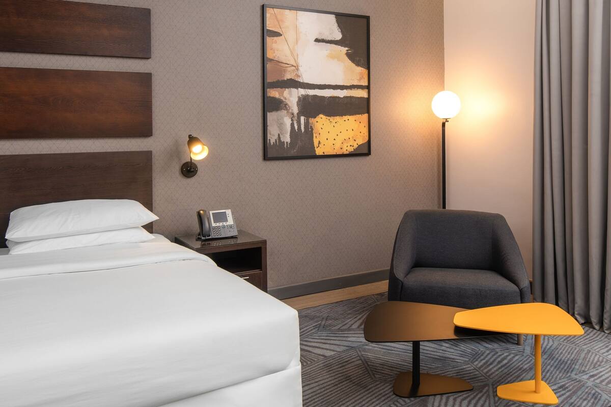 A hotel room with a bed, a chair, a small table, a wall-mounted phone, and a painting.