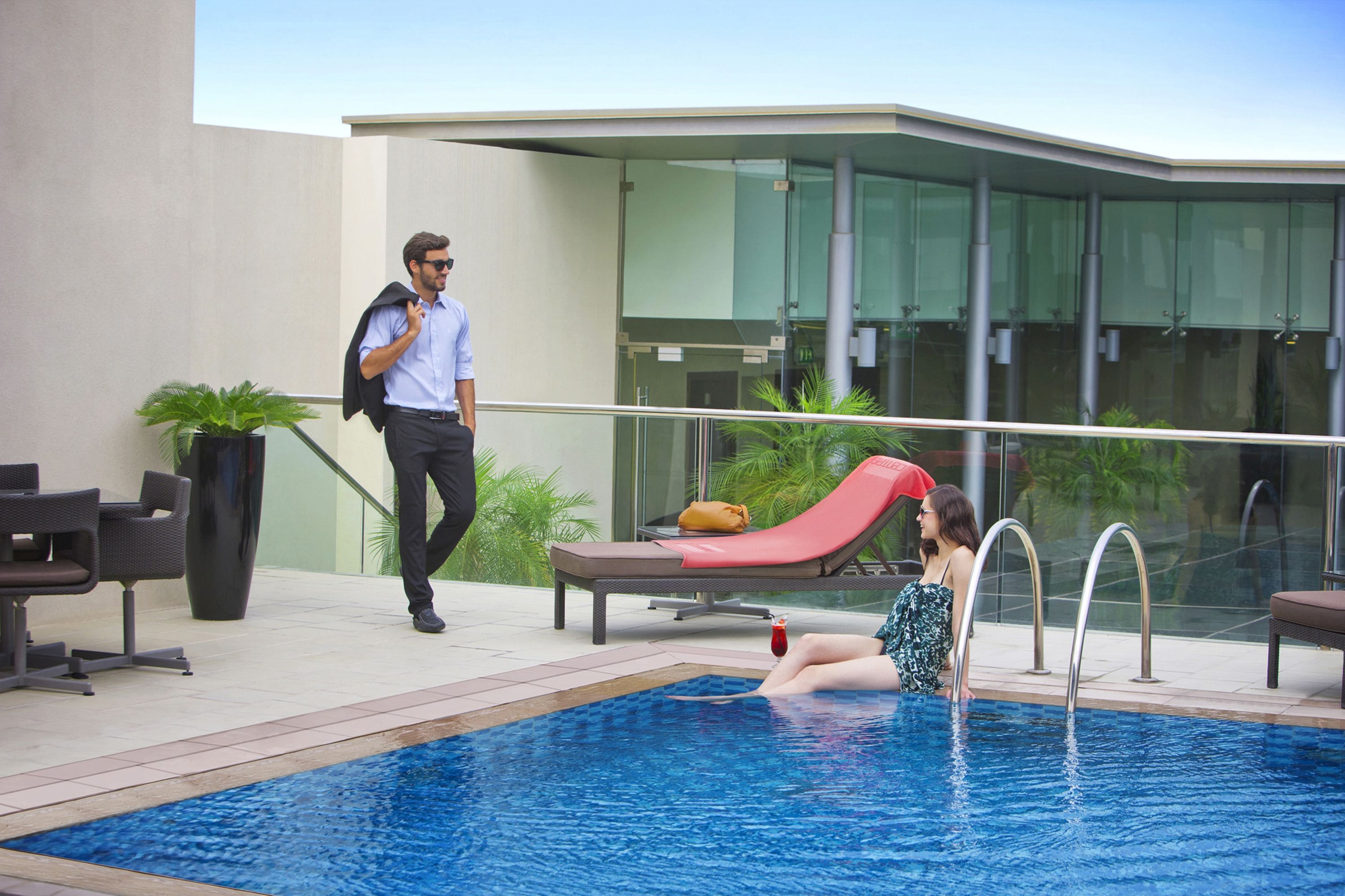 Un espace piscine moderne dans un hôtel, avec une balustrade en verre, des chaises longues et une chaise longue rouge.