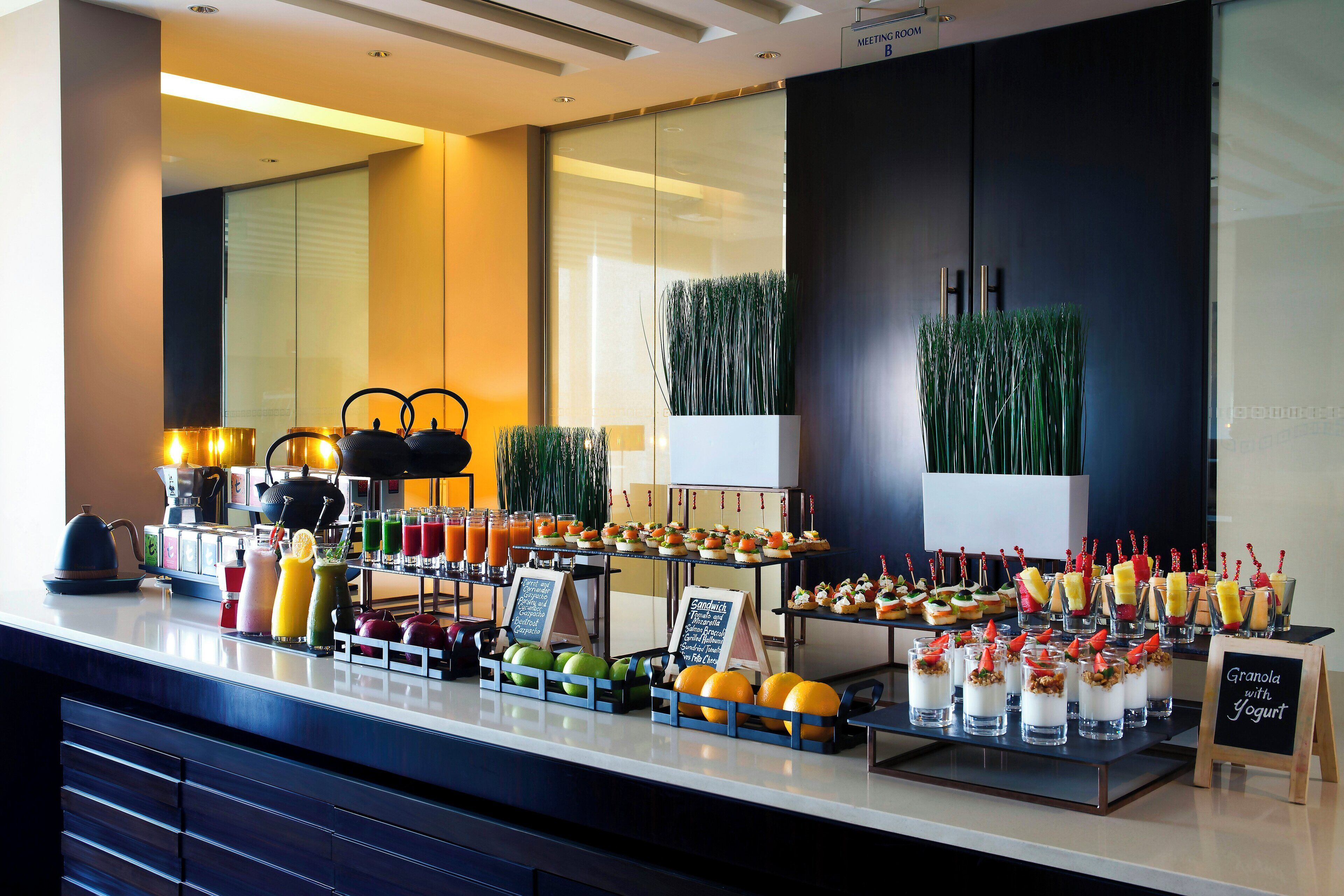 A buffet with various food items and drinks, a meeting room sign, and indoor plants.