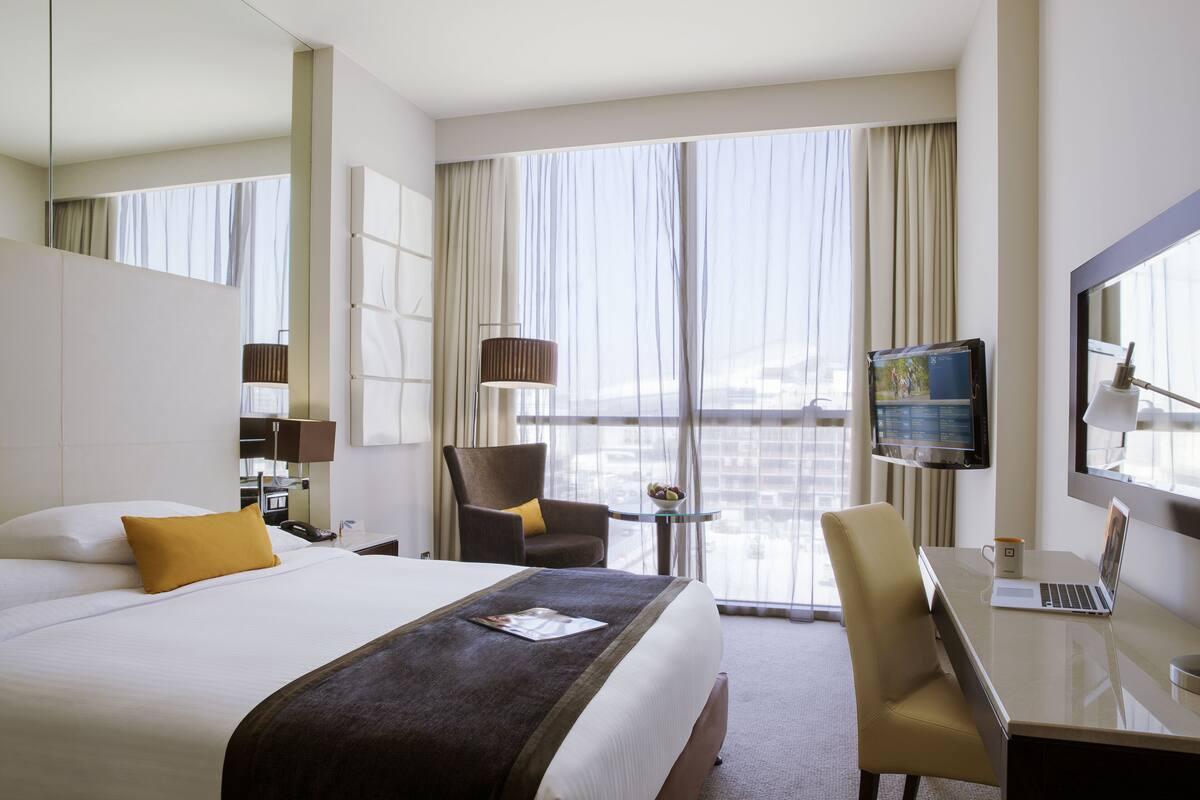 A hotel room with a large bed, a desk with a laptop, a chair, a TV, and a balcony with curtains.