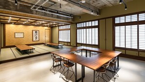 Ruime kamer met een grote houten tafel en stoelen, grote ramen met een traditioneel kruisraamontwerp, en een betonplafond met zichtbare leidingen.