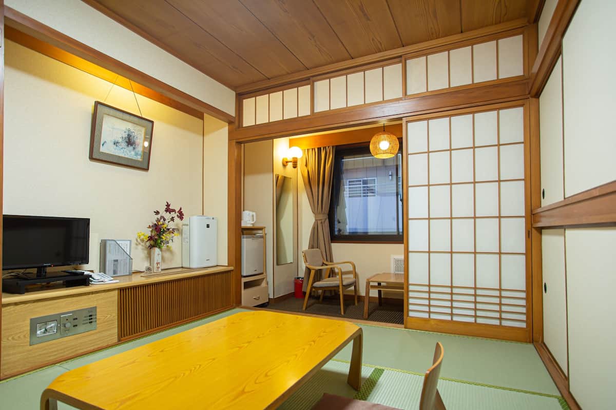 A traditional Japanese-style room with tatami flooring, a wooden table, a television, a refrigerator, and a window with curtains.