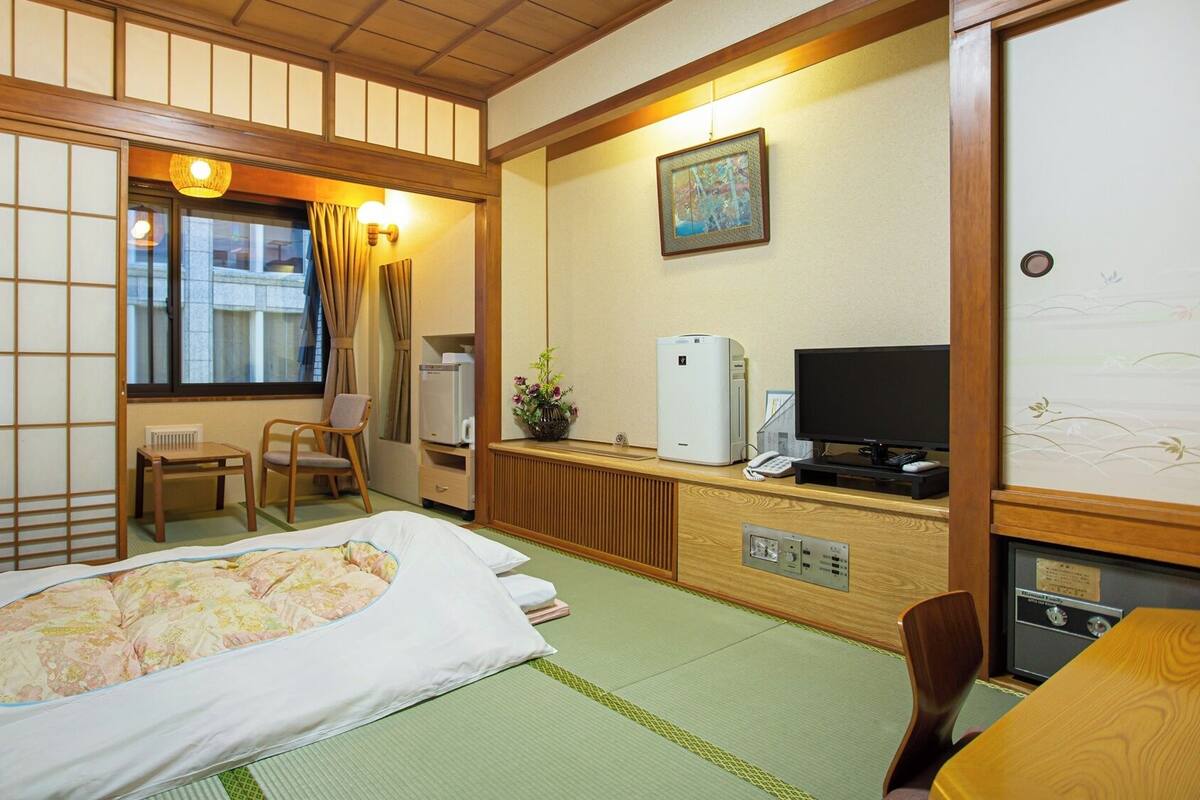 A traditional Japanese room with tatami flooring, a bed, a desk with a computer, a television, and a window with curtains.