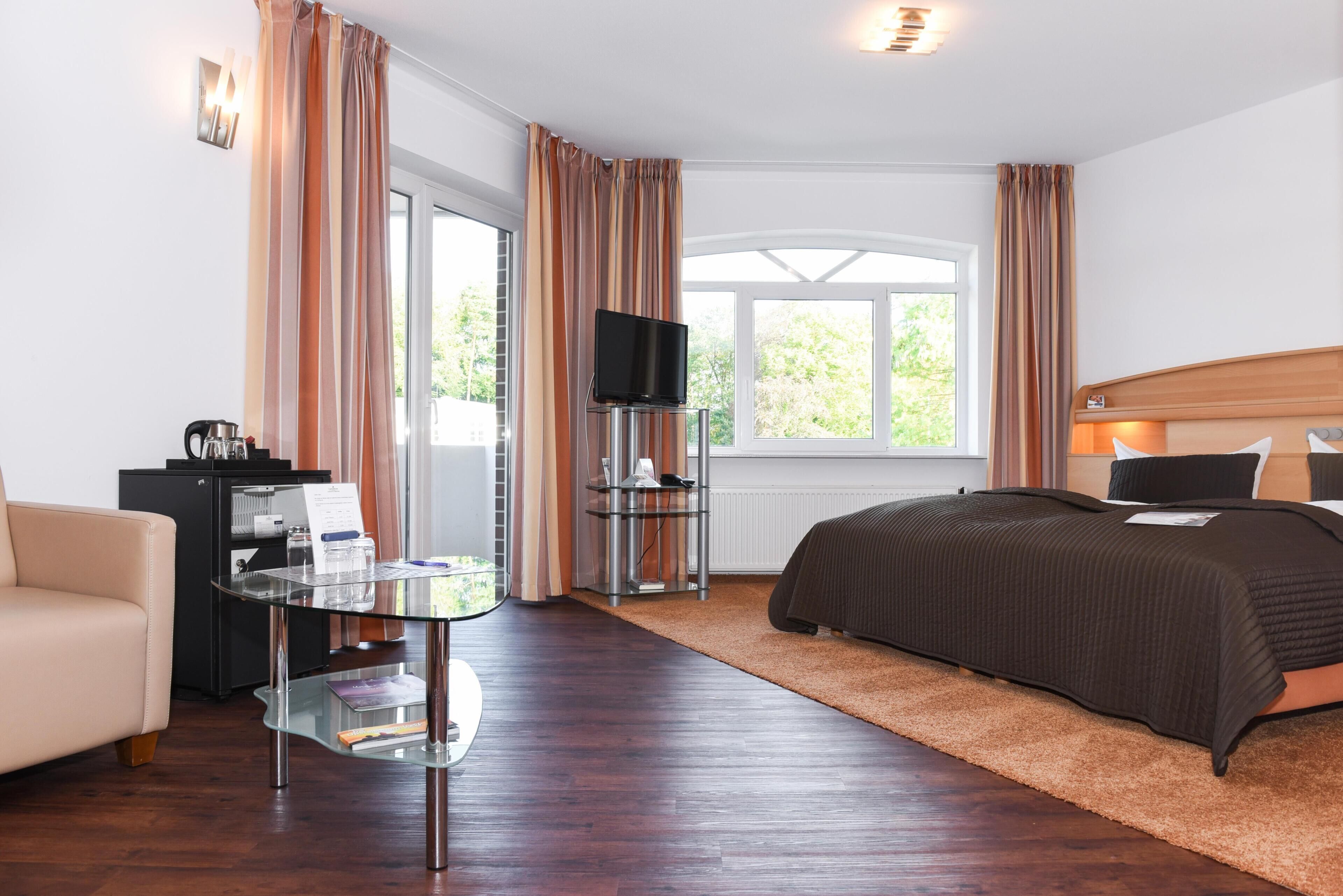 Habitación de hotel con cama, televisor, mesa de vidrio, sofá y ventana con cortinas.