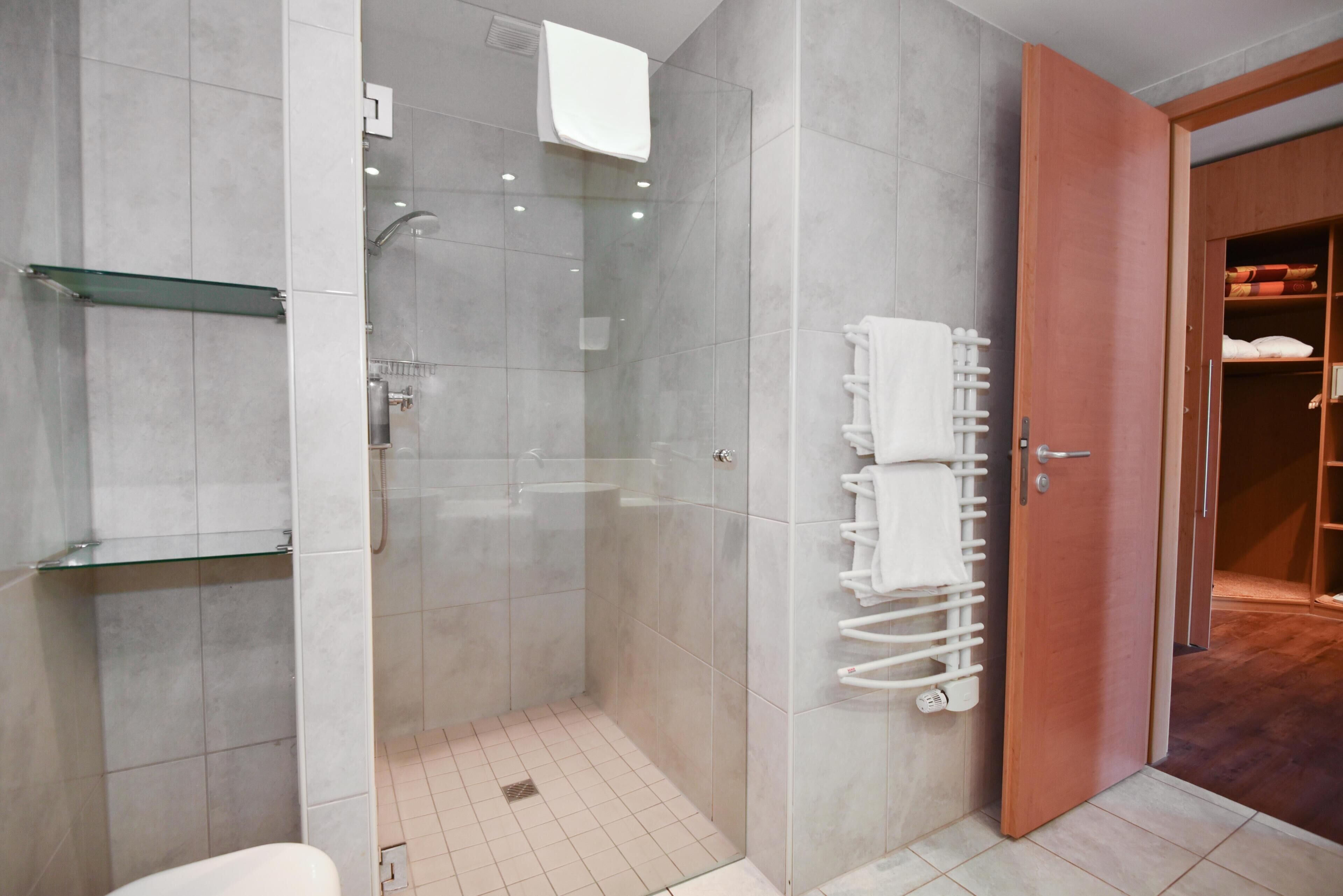 Un baño moderno con ducha envuelta en vidrio, repisa de vidrio, perchero y puerta de madera que da a un armario.