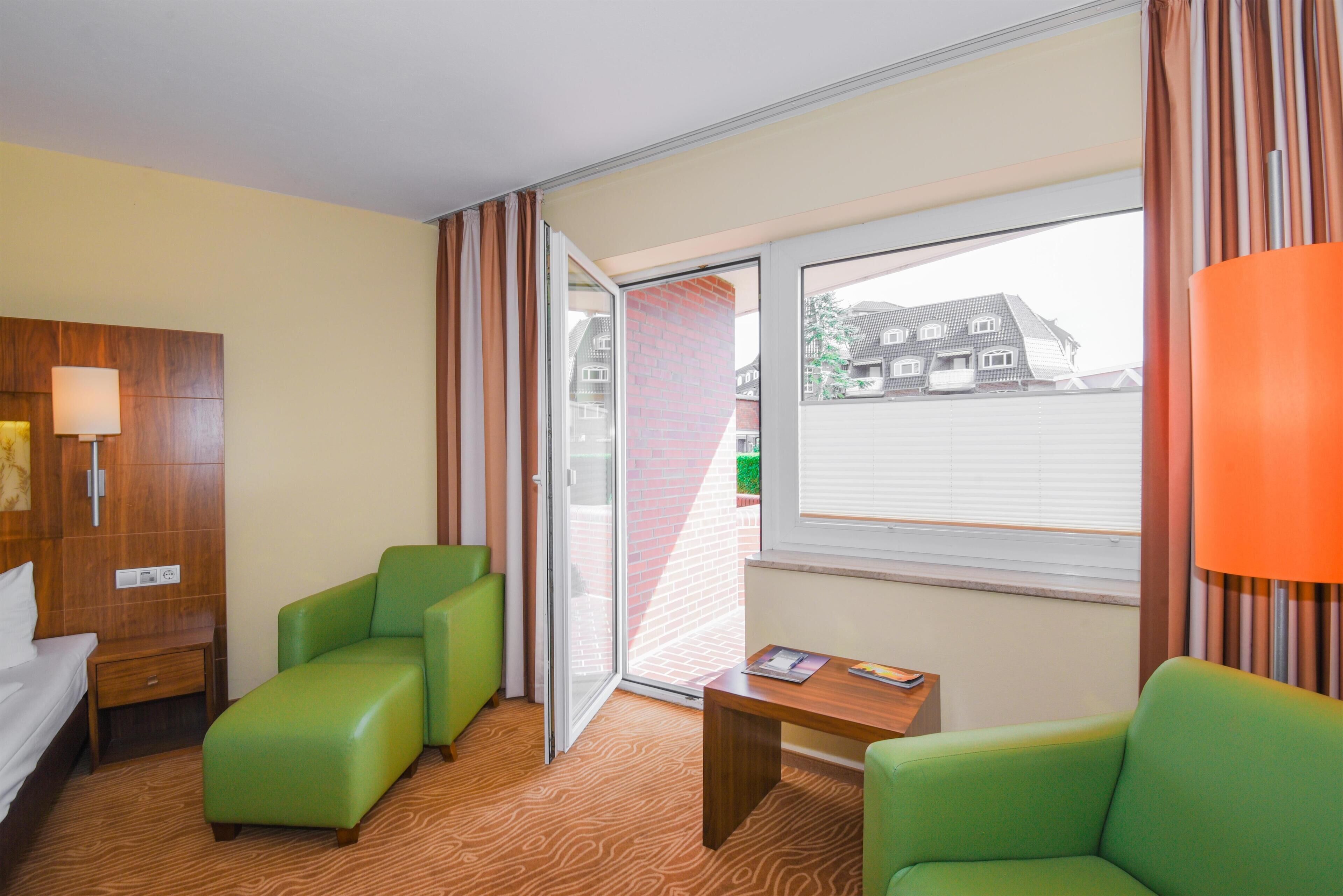 Ein Hotelzimmer mit Bett, grünen Sesseln, einem Holztisch, einer Lampe und einem Fenster mit Vorhängen.