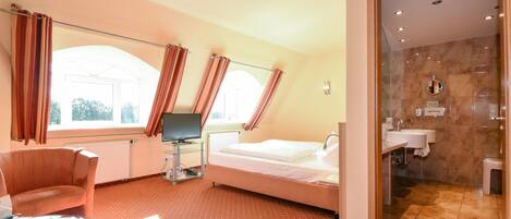 A hotel room with a bed, a desk with a computer, a chair, a glass table, a bathroom with a sink and mirror, and large windows with curtains.