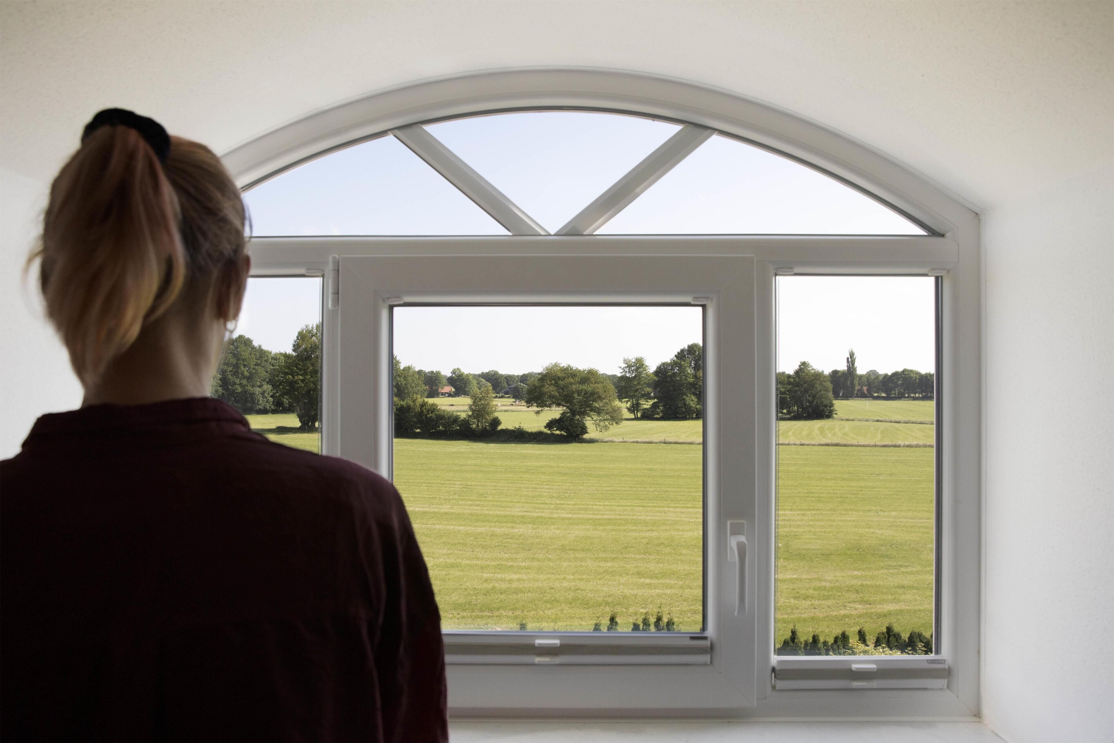 Uma pessoa em pé num quarto com uma janela grande e arqueada com vista para um campo verde.