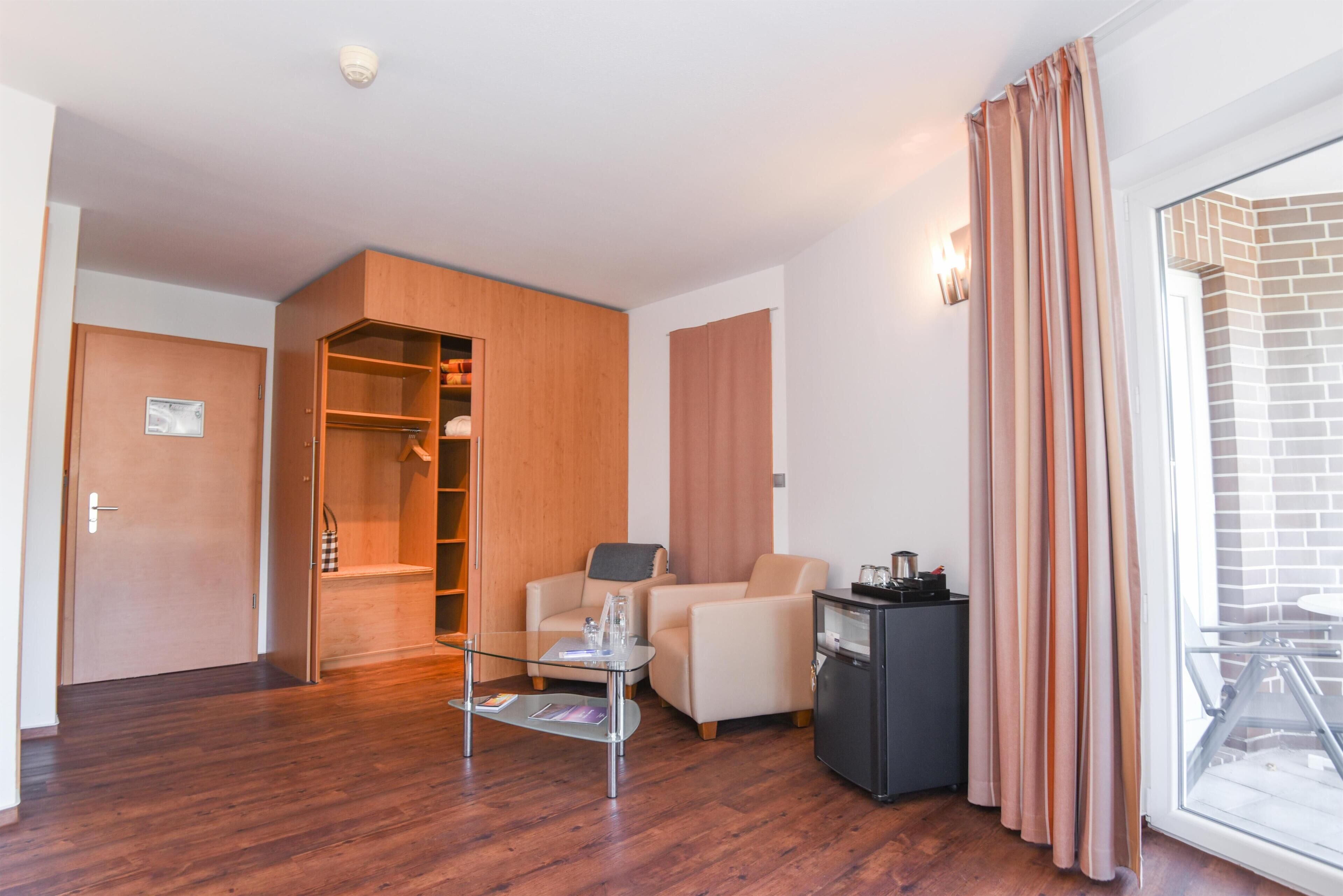 A modern hotel room with a wooden wardrobe, a seating area with a glass table, and a view through a glass door.